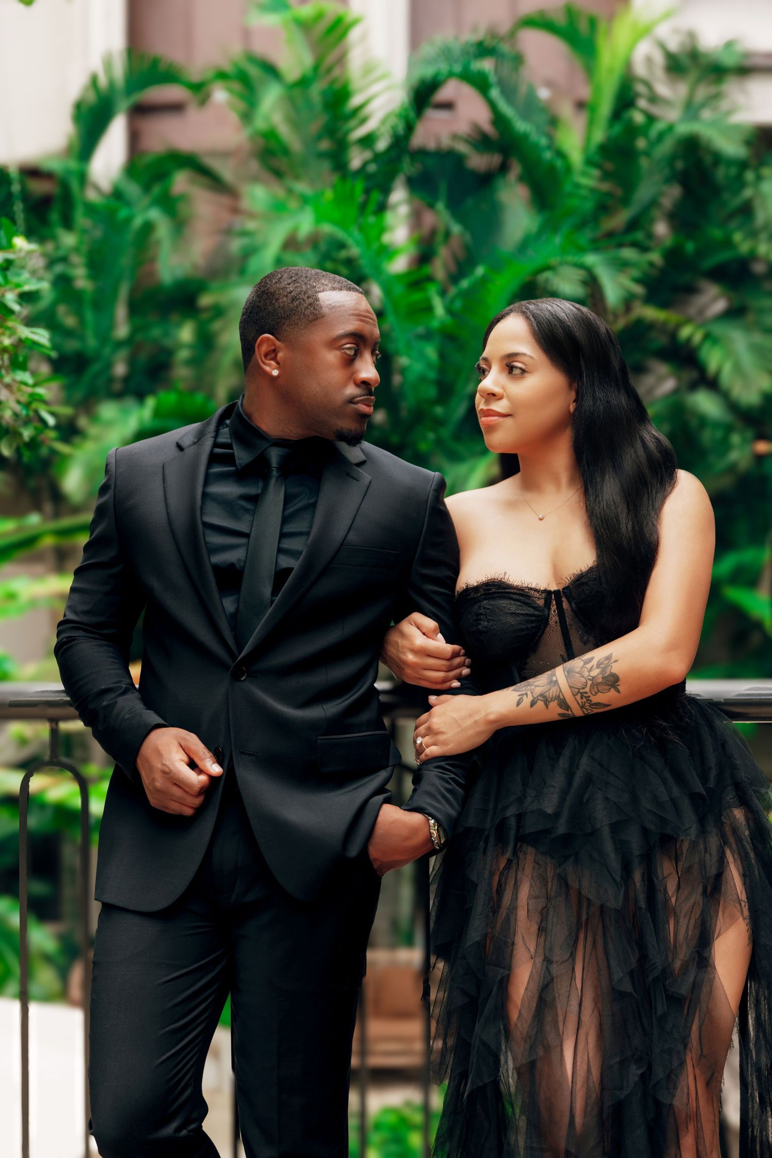Couple in formal black attire posing together against tropical greenery backdrop.