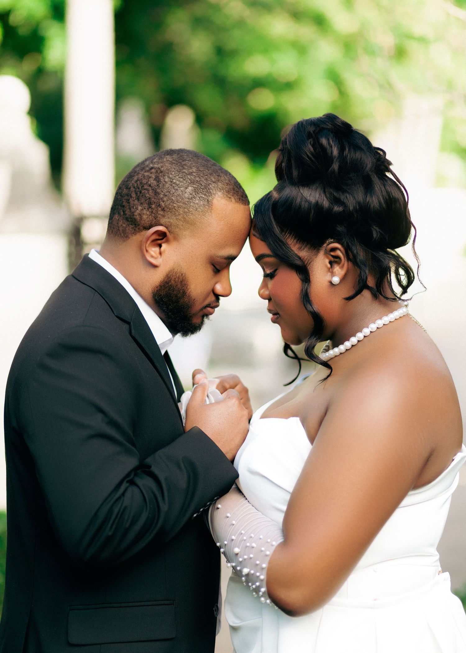A bride and groom share an intimate forehead touch moment outdoors, dressed in wedding attire with pearls and a black tuxedo.