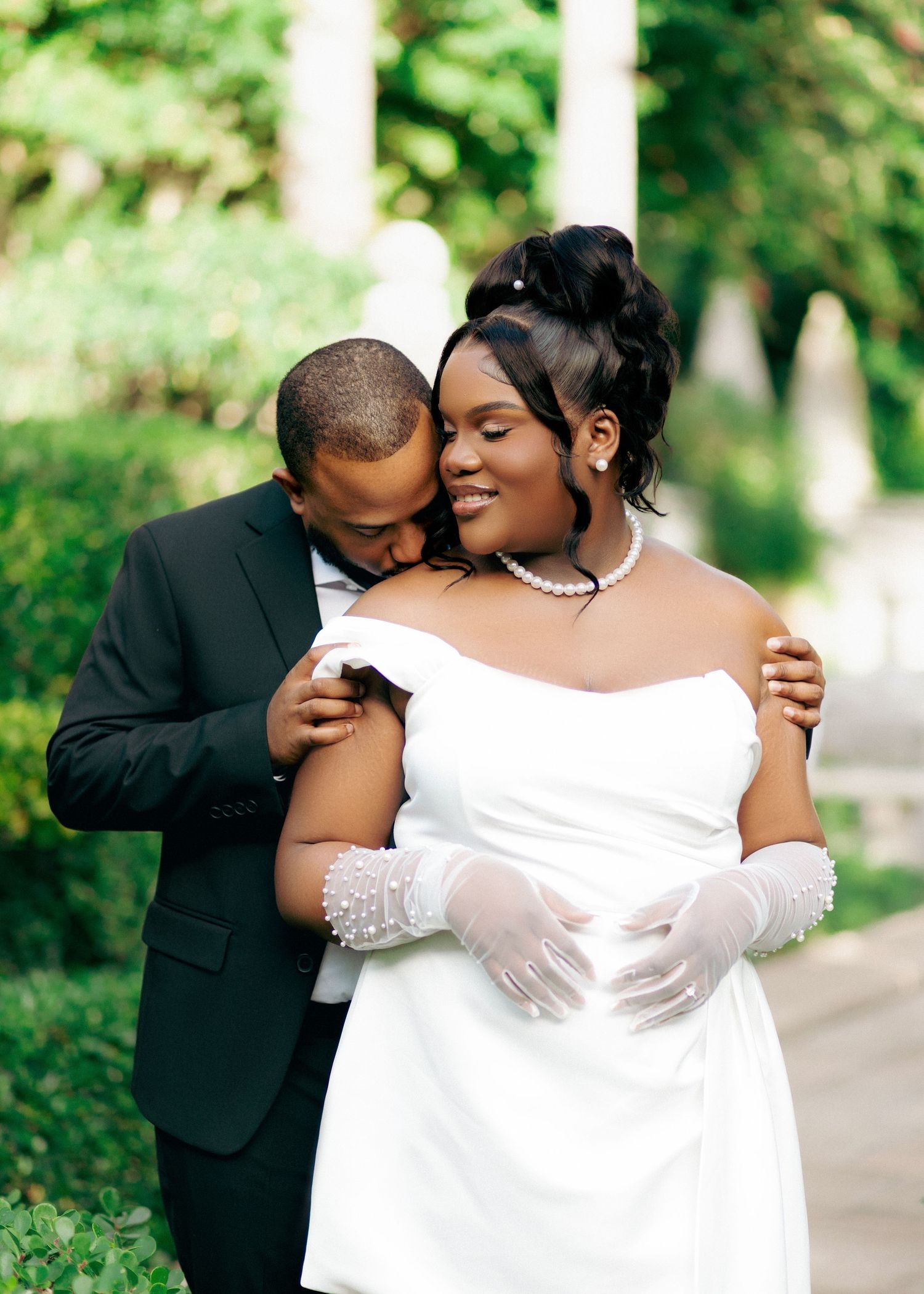 A romantic outdoor wedding portrait showing a couple in formal attire sharing an intimate moment among greenery.