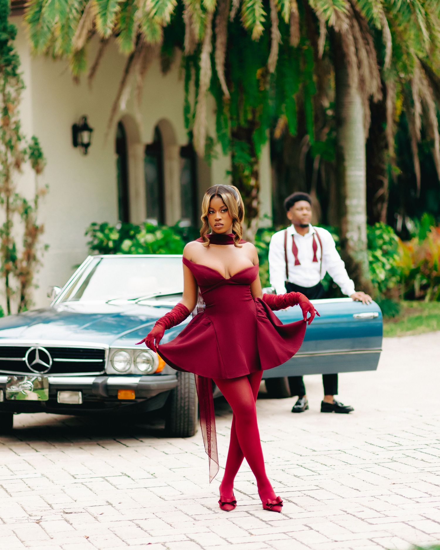 Stylish figure in burgundy dress and tights poses by vintage Mercedes-Benz car in tropical courtyard setting.