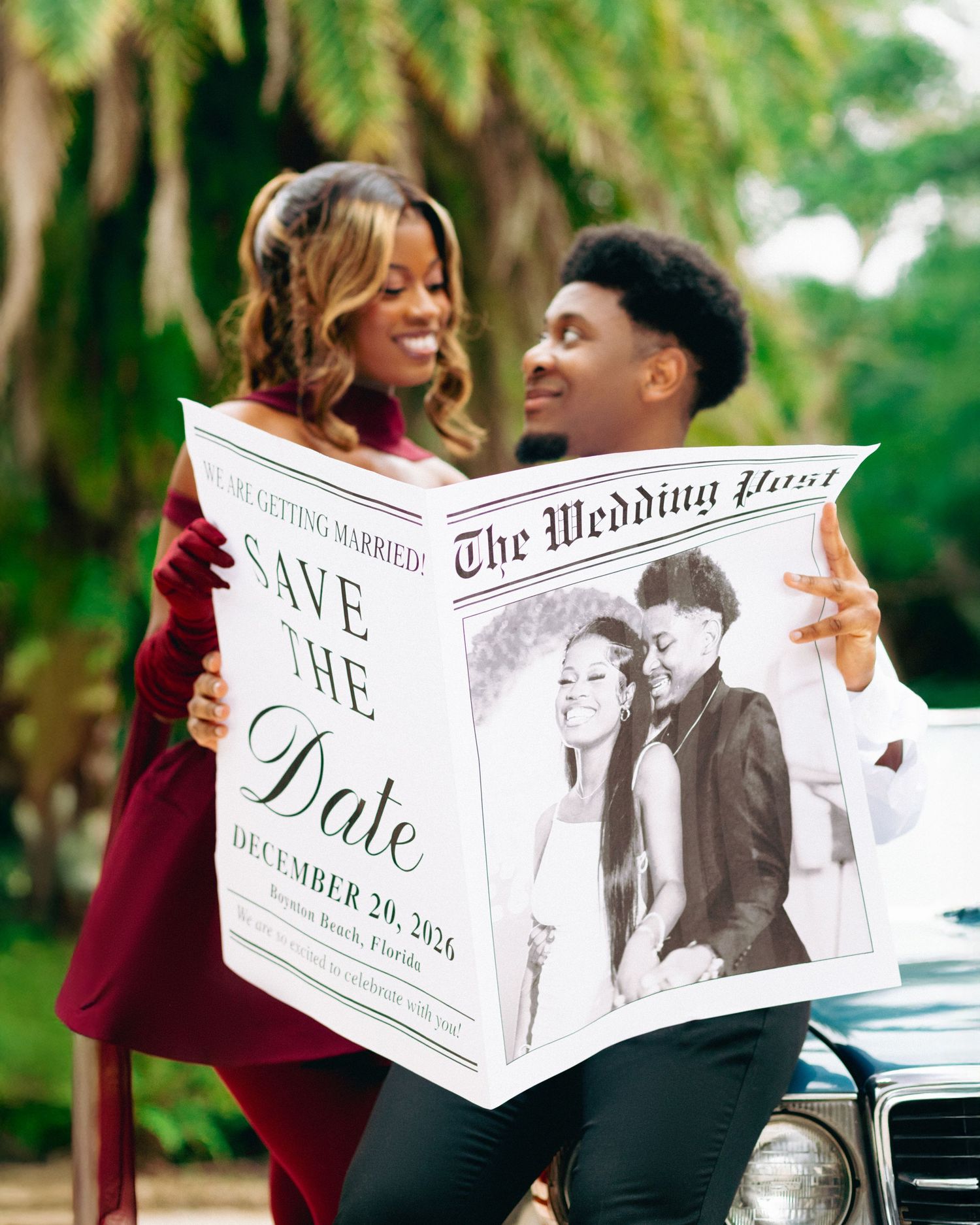 A couple holds up a stylized newspaper-themed save the date announcement in an outdoor setting with palm trees.