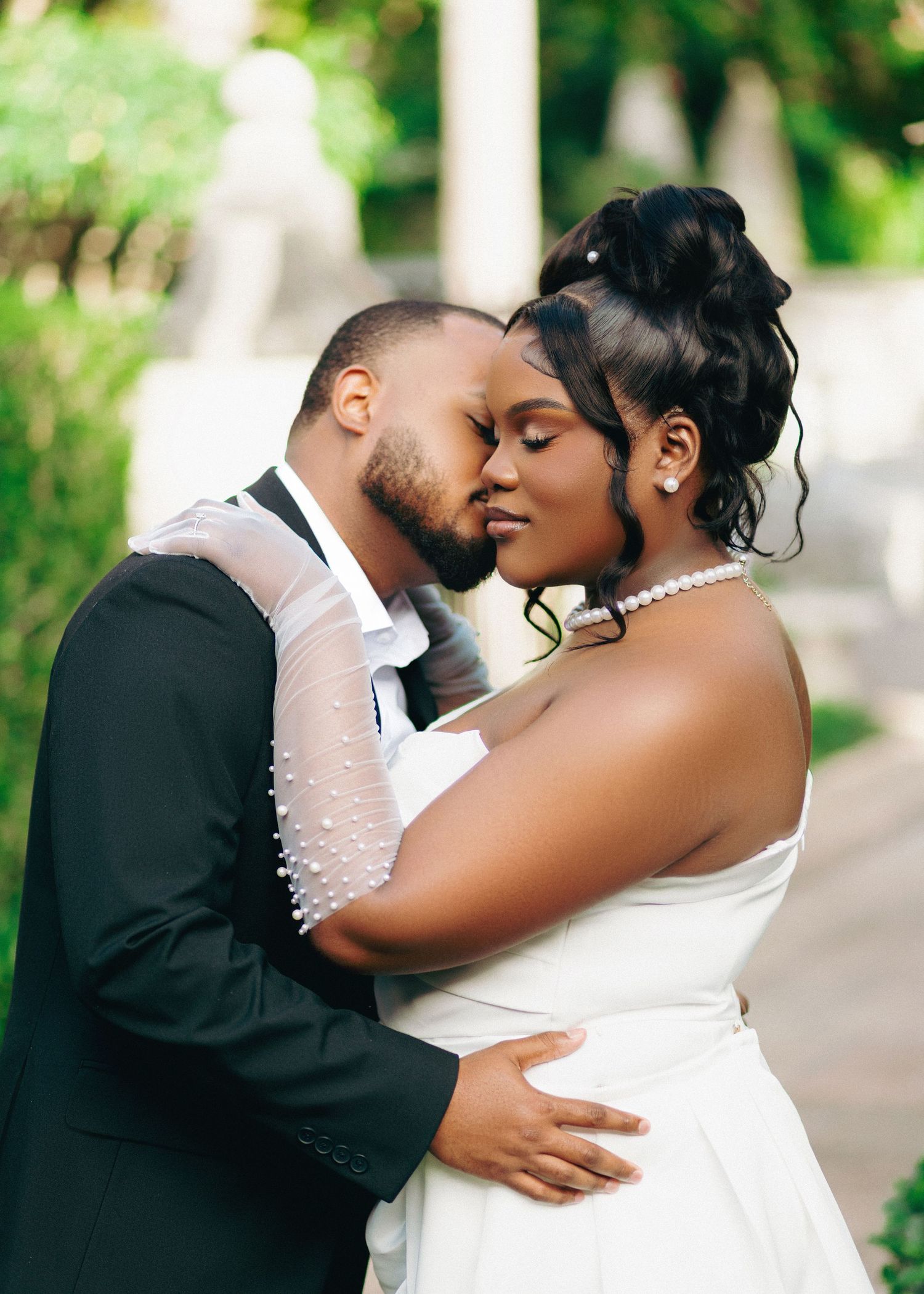 A bride and groom share a romantic kiss outdoors, dressed in wedding attire with lush green trees in the background.