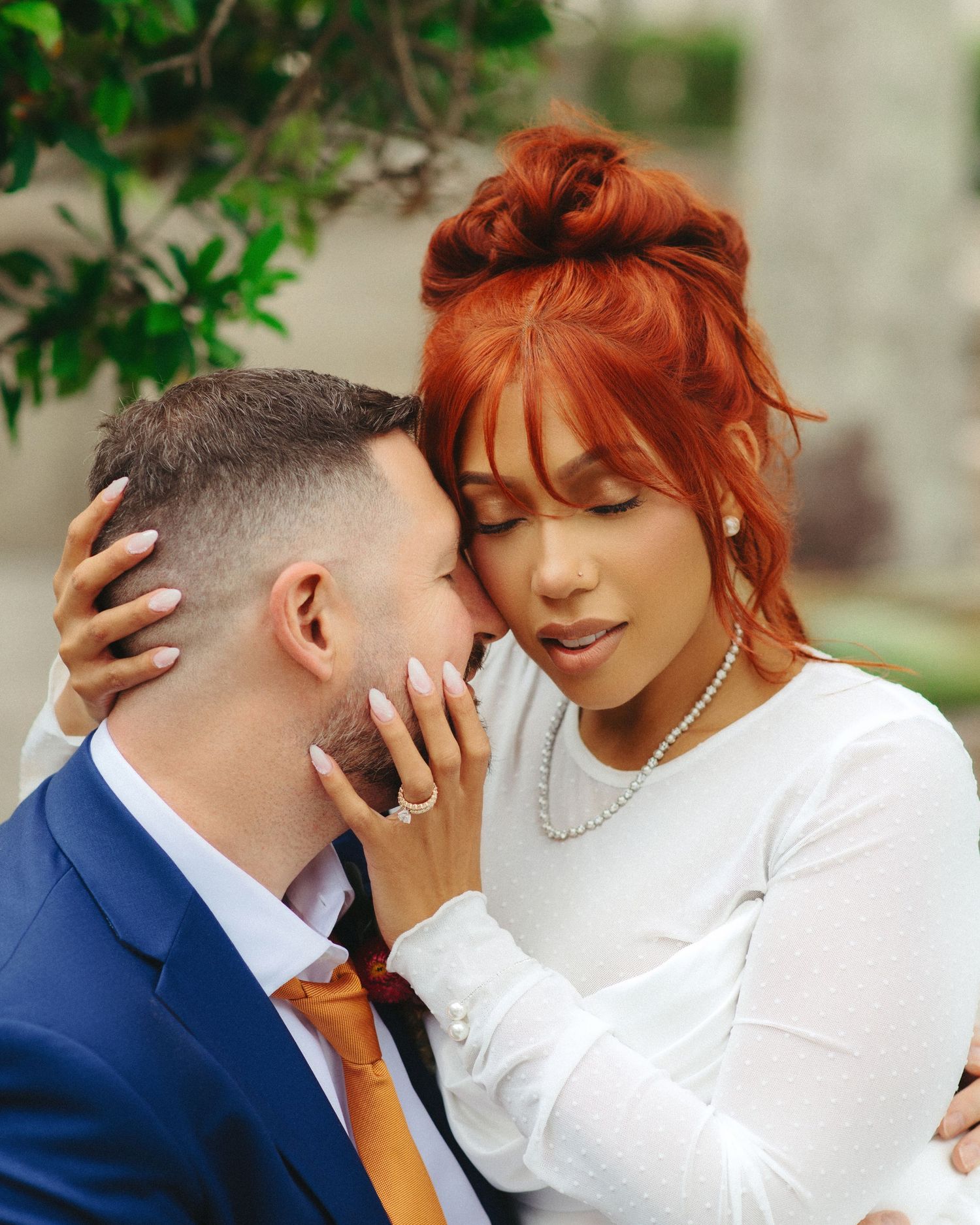 A couple shares an intimate moment outdoors, with one wearing a navy suit and orange tie and the other in white.