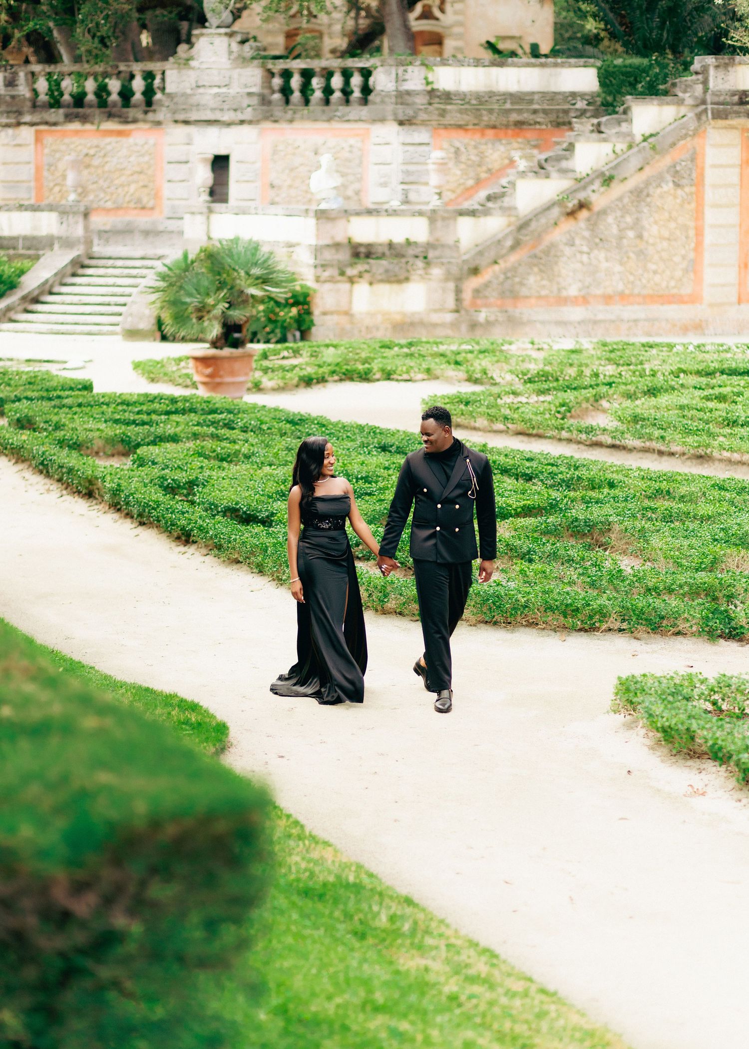 Two people walking along a curved garden path surrounded by manicured hedges and historic stone architecture.