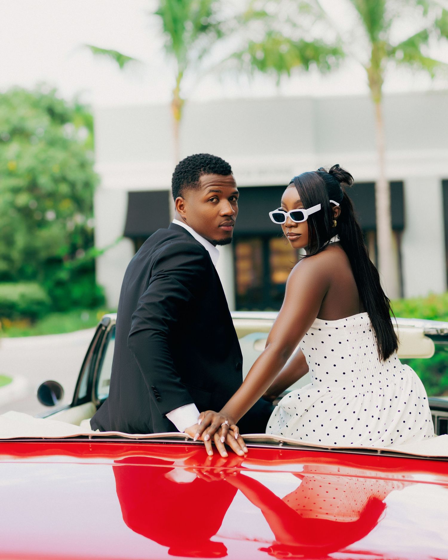 Romantic couple posing together with a classic red convertible car in a tropical setting.