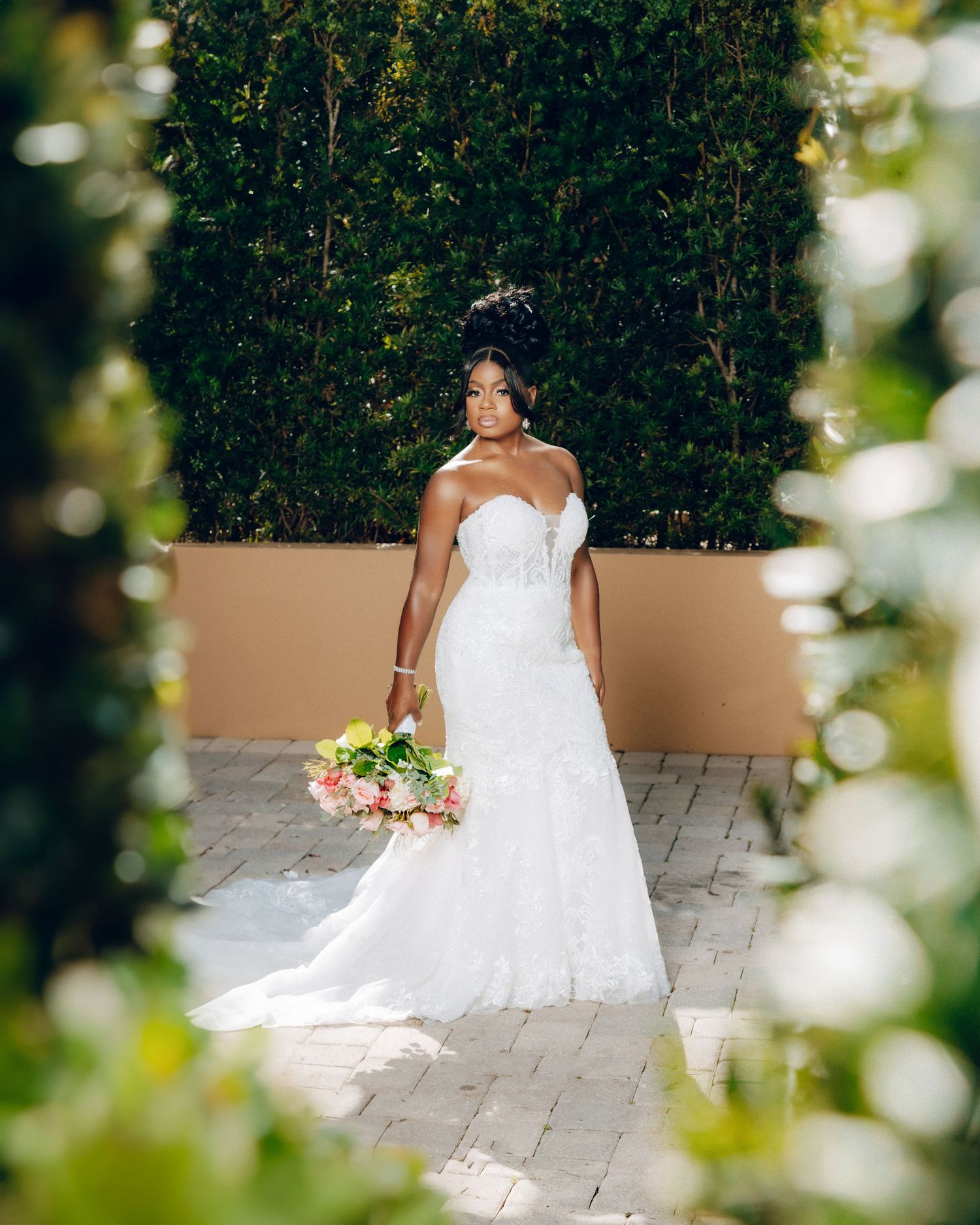 Bride in white lace gown poses gracefully on stone pathway surrounded by flowers.