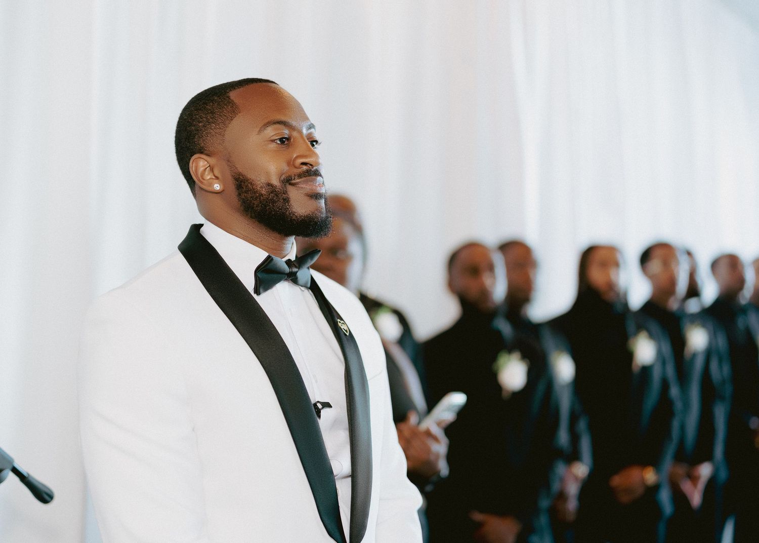 Groom in white tuxedo smiling at wedding ceremony with groomsmen in background.