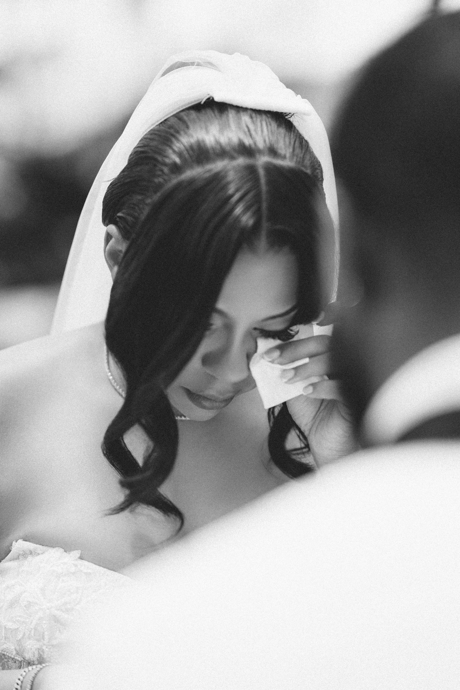 Black and white photo of a bride wiping tears during wedding ceremony, wearing veil and lace dress.
