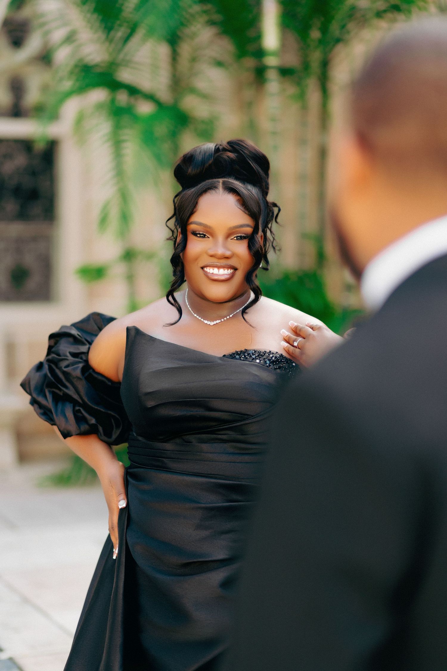 A smiling woman in an elegant black off-shoulder gown with ruffle sleeves and pearl necklace at a formal event.