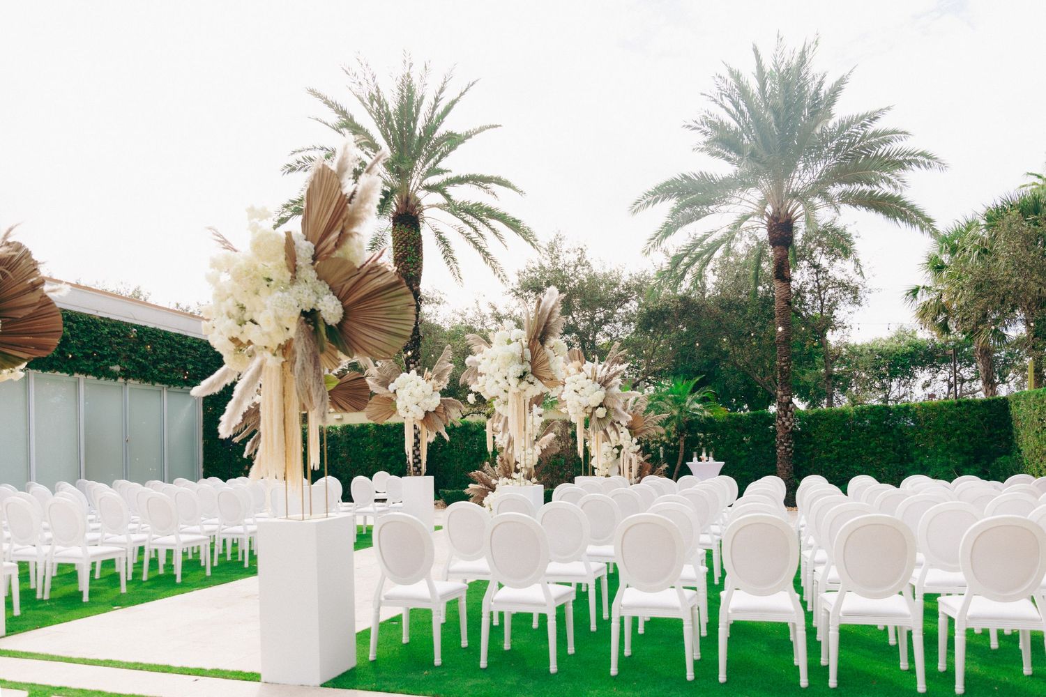 Elegant outdoor wedding venue setup with white chairs, palm trees, and decorative bows on a green lawn.