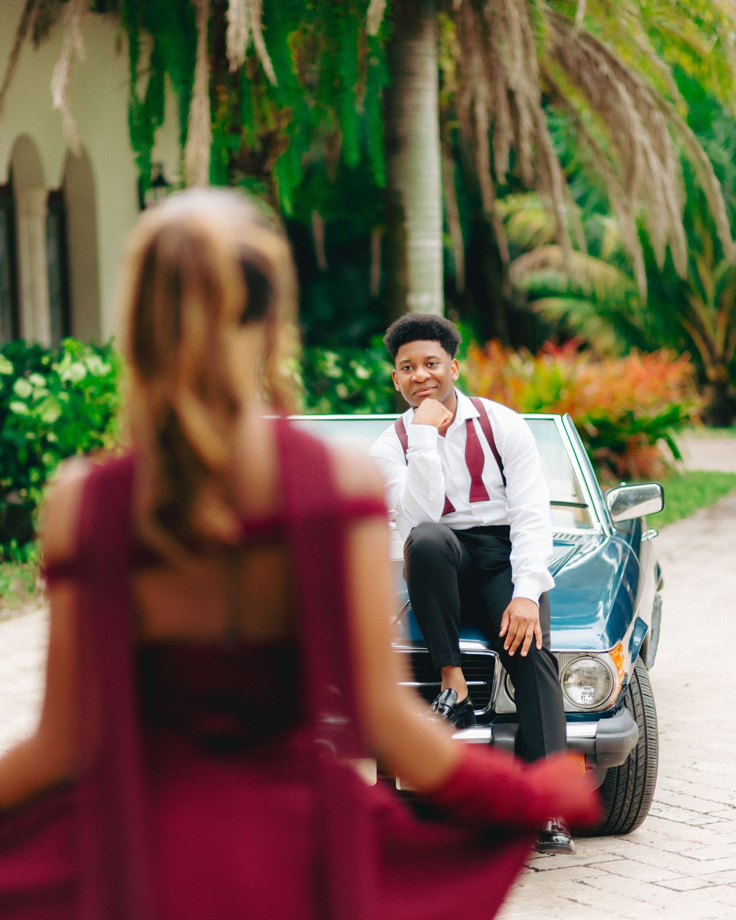 A vintage car scene with someone in burgundy dress in foreground and another person sitting on classic car in background.