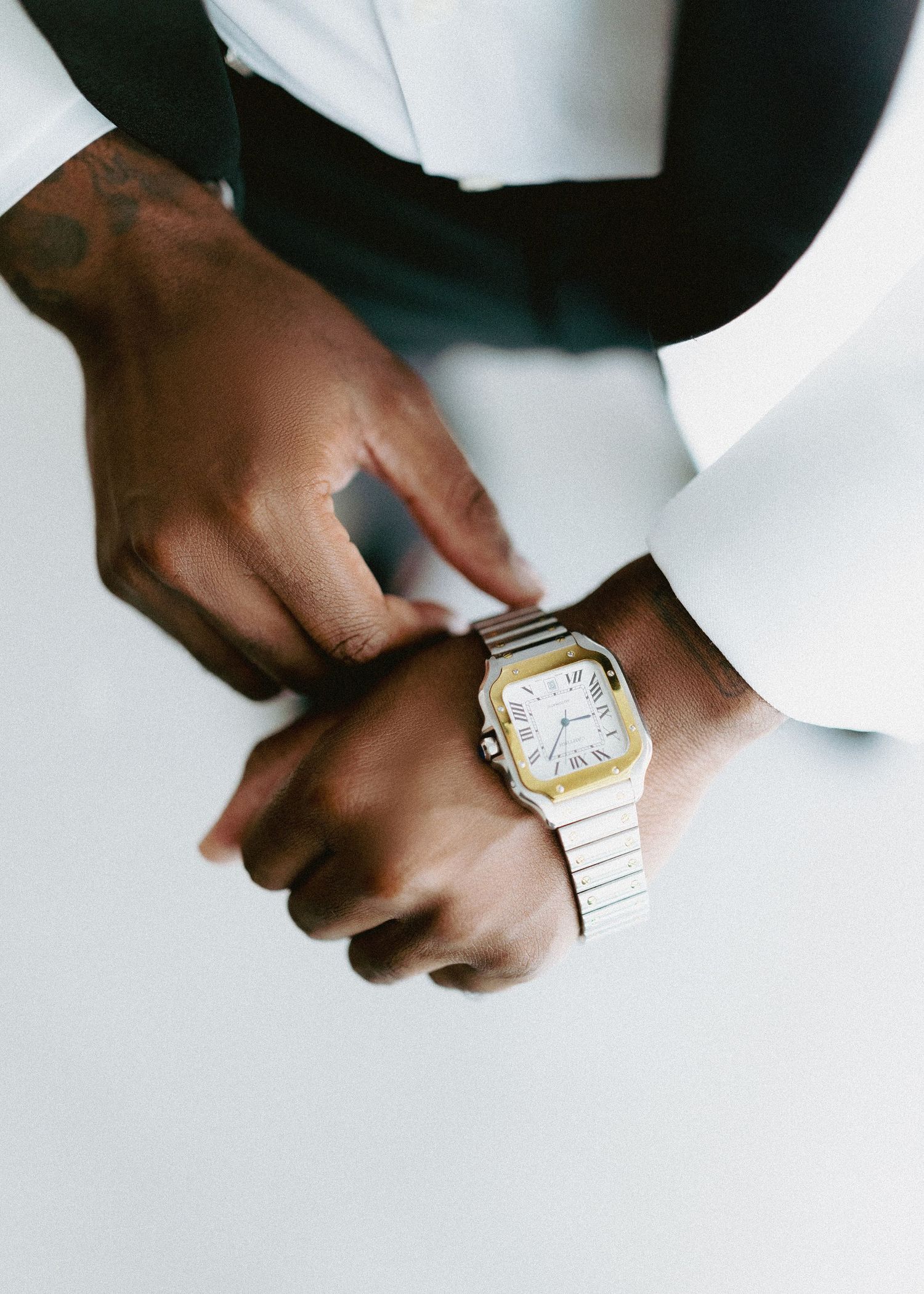 Groom adjusting gold and silver Cartier Santos watch on his wrist, wearing white tuxedo jacket.