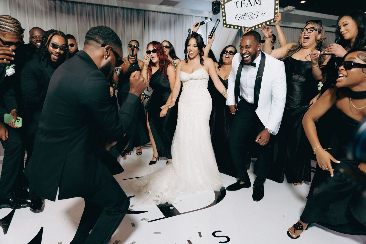 Bride and groom dance joyfully with wedding guests on white dance floor at reception.