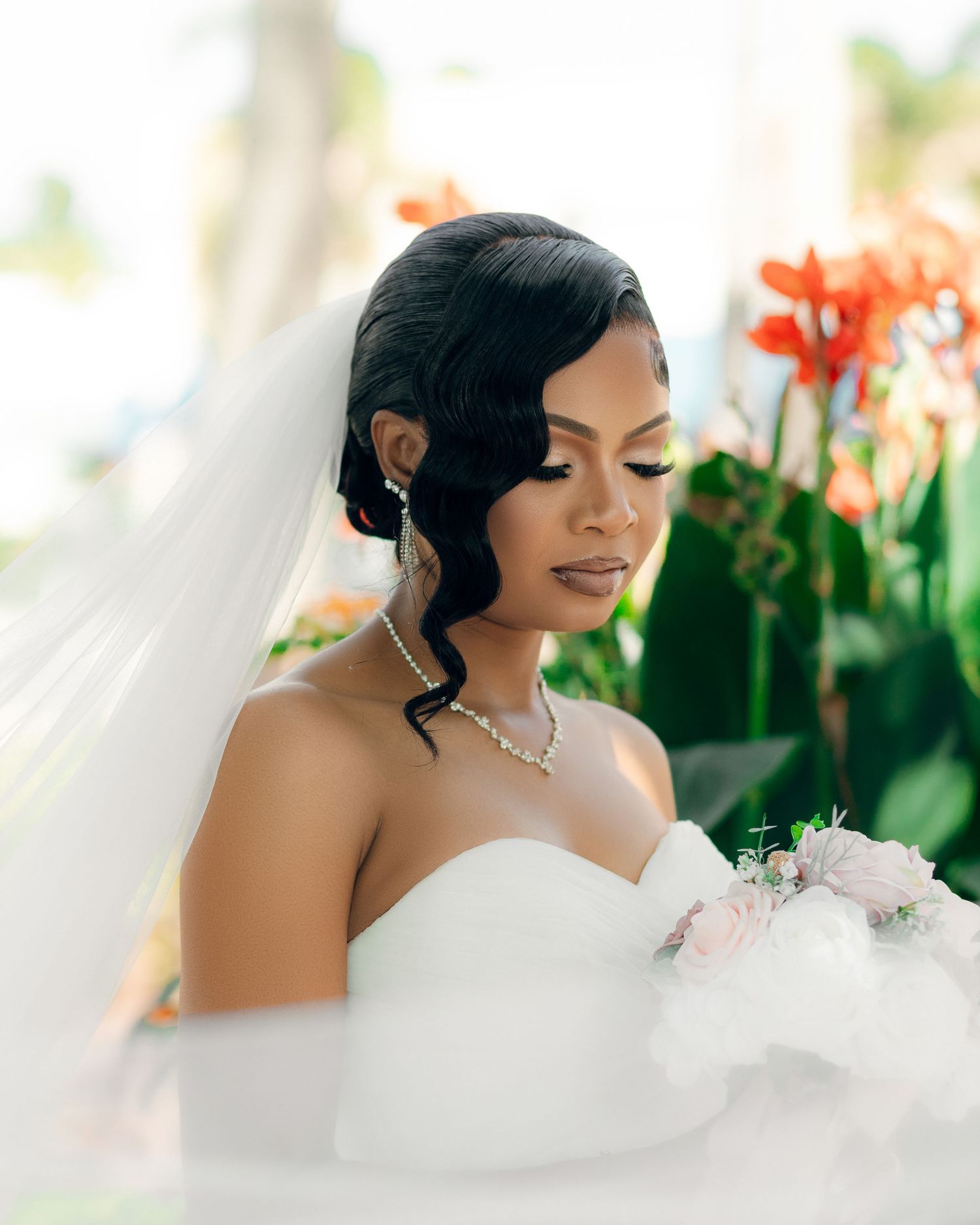 A bride in a white strapless gown and elegant jewelry poses with orange lilies and greenery in the background.