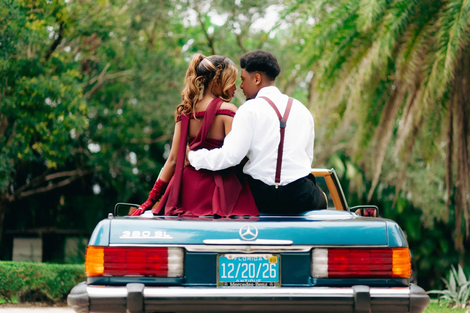 Couple sitting on back of vintage convertible car with palm trees in background wearing burgundy dress and suspenders.