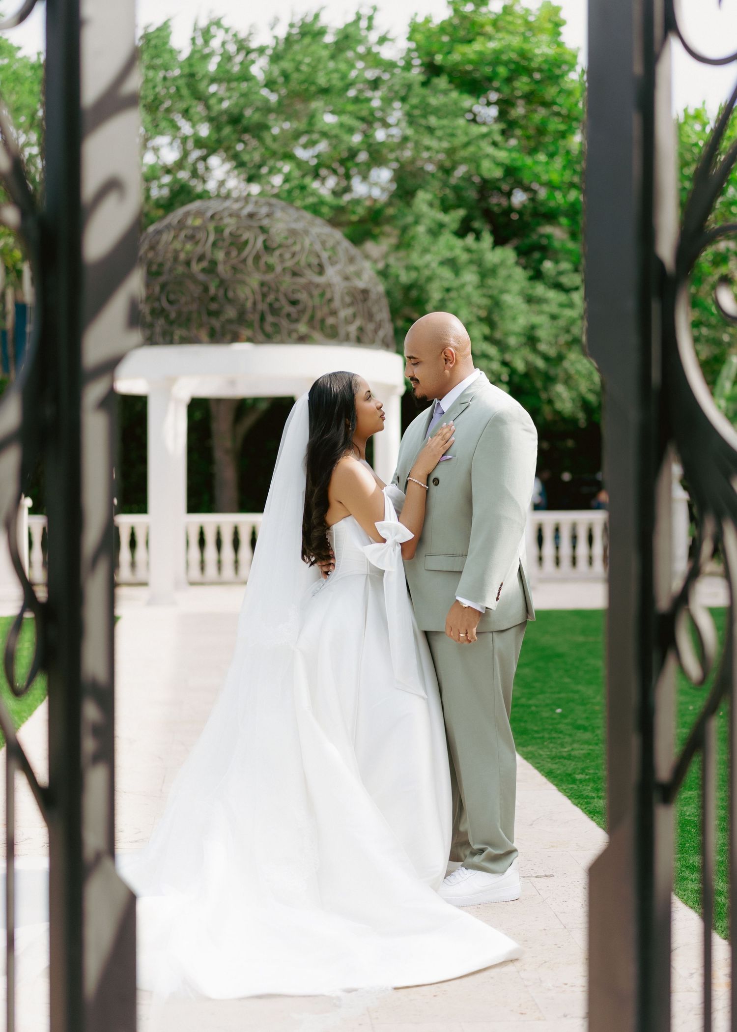 Bride in white gown and veil stands with groom in gray suit framed by iron gates at elegant outdoor wedding venue.