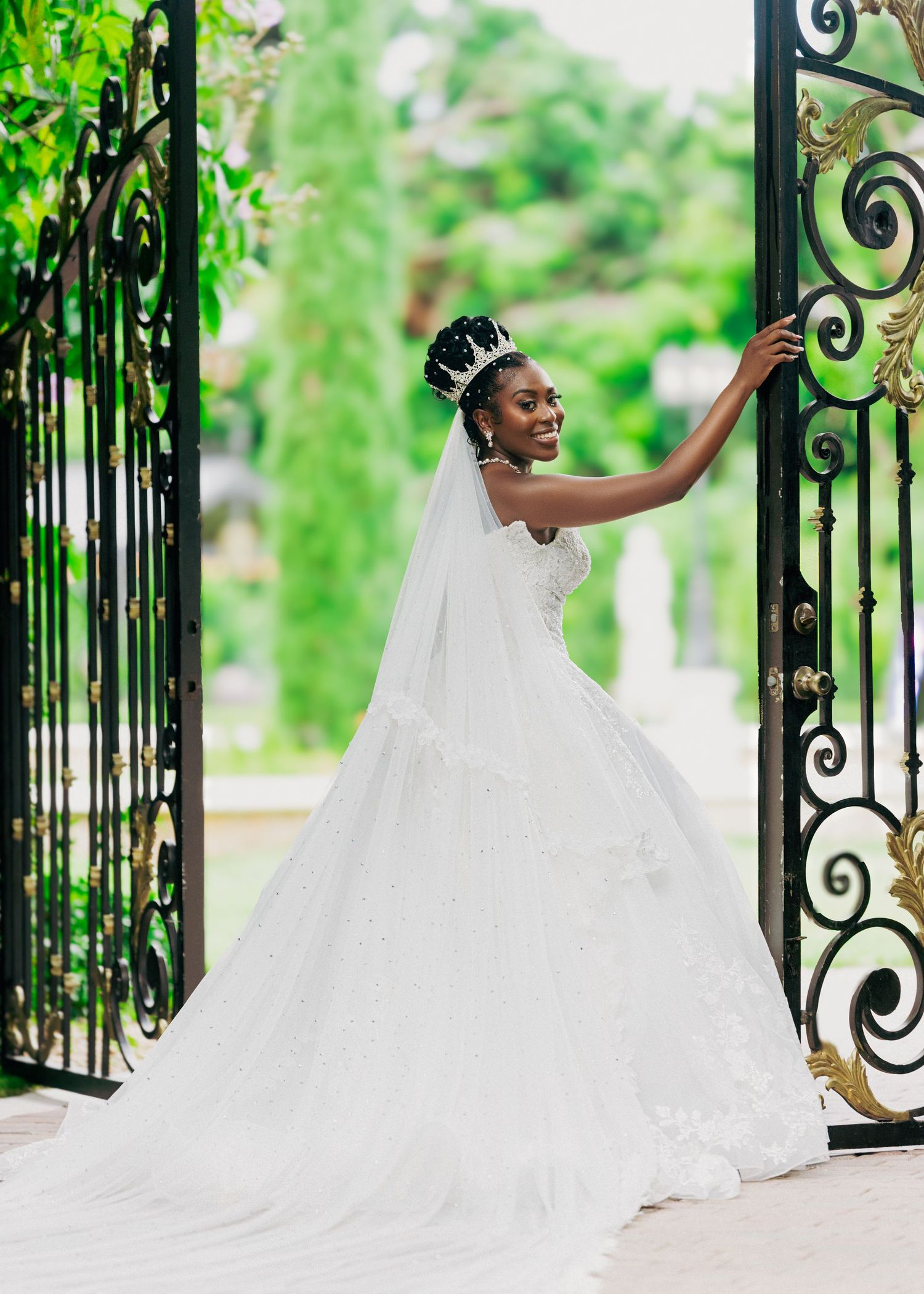 Bride in flowing white gown poses along ornate iron gates with lush greenery backdrop.