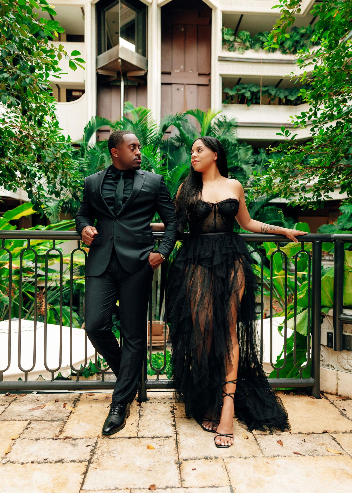 Romantic couple in black formal attire poses together against lush greenery in an outdoor setting.