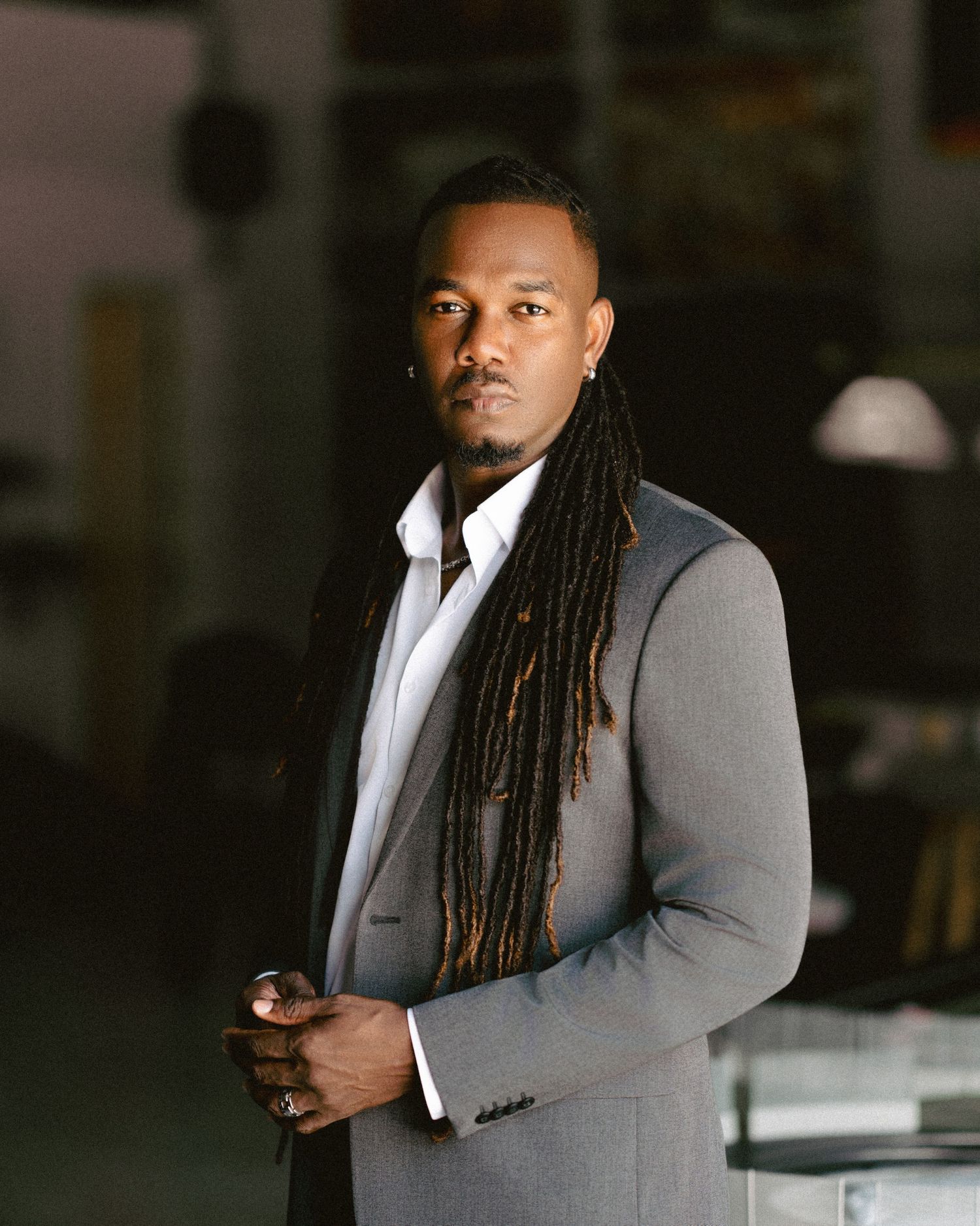 A well-dressed man in a gray suit with long dreadlocks poses confidently in a stylish indoor setting.