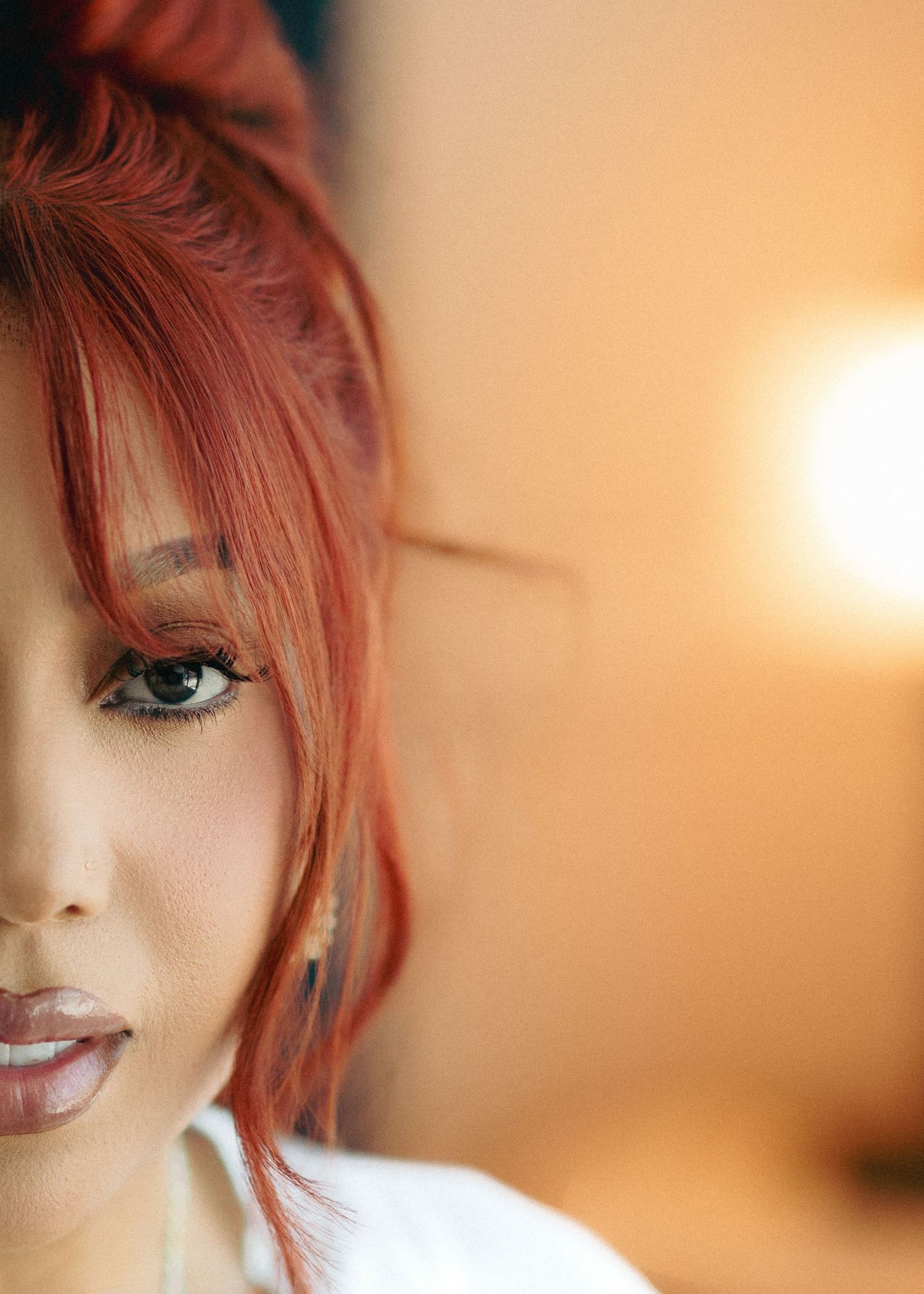 Close up portrait with bright red hair and dramatic eye makeup against a warm peachy background.