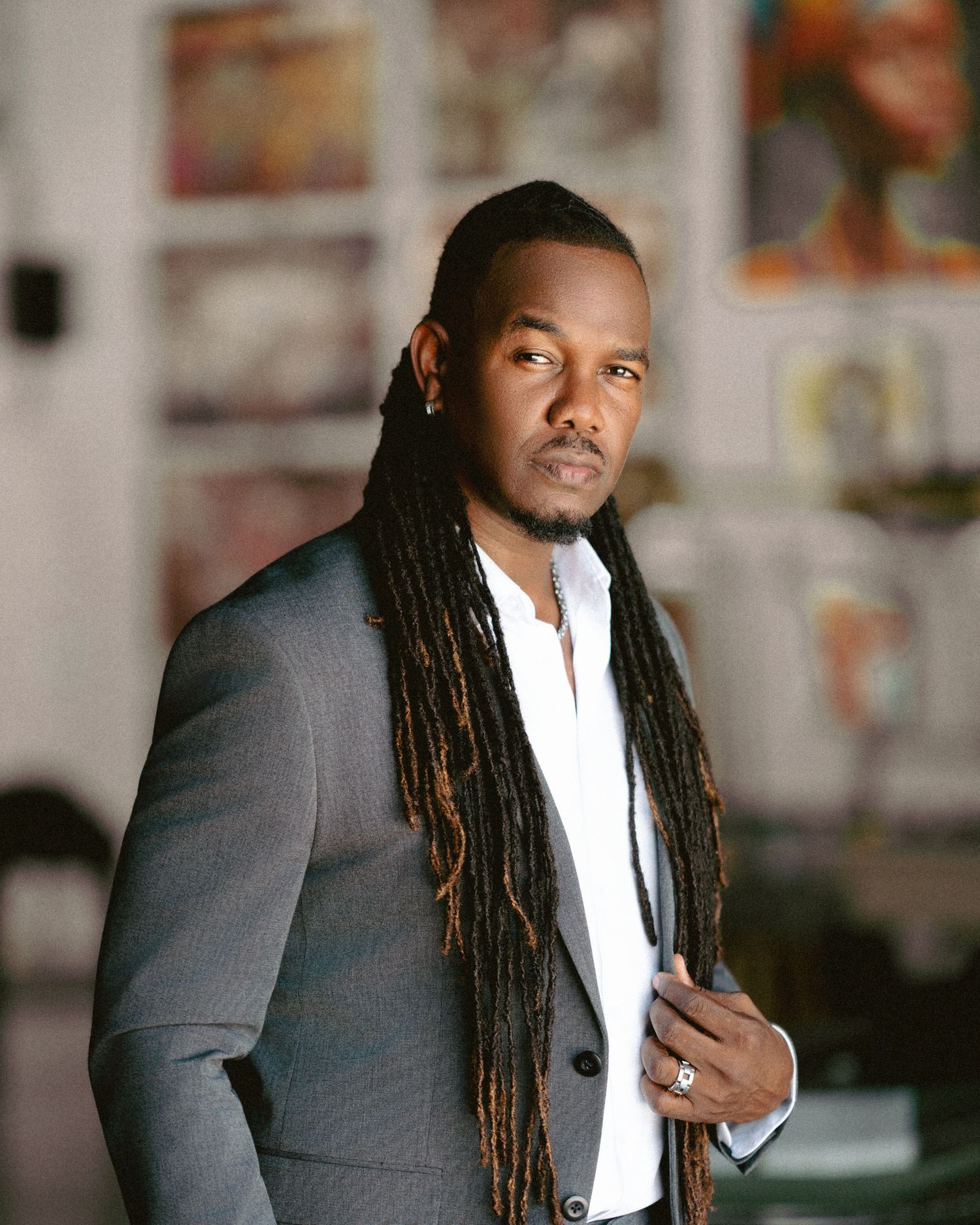 Man with long dreadlocks wearing a gray suit poses confidently indoors with colorful artwork in background.