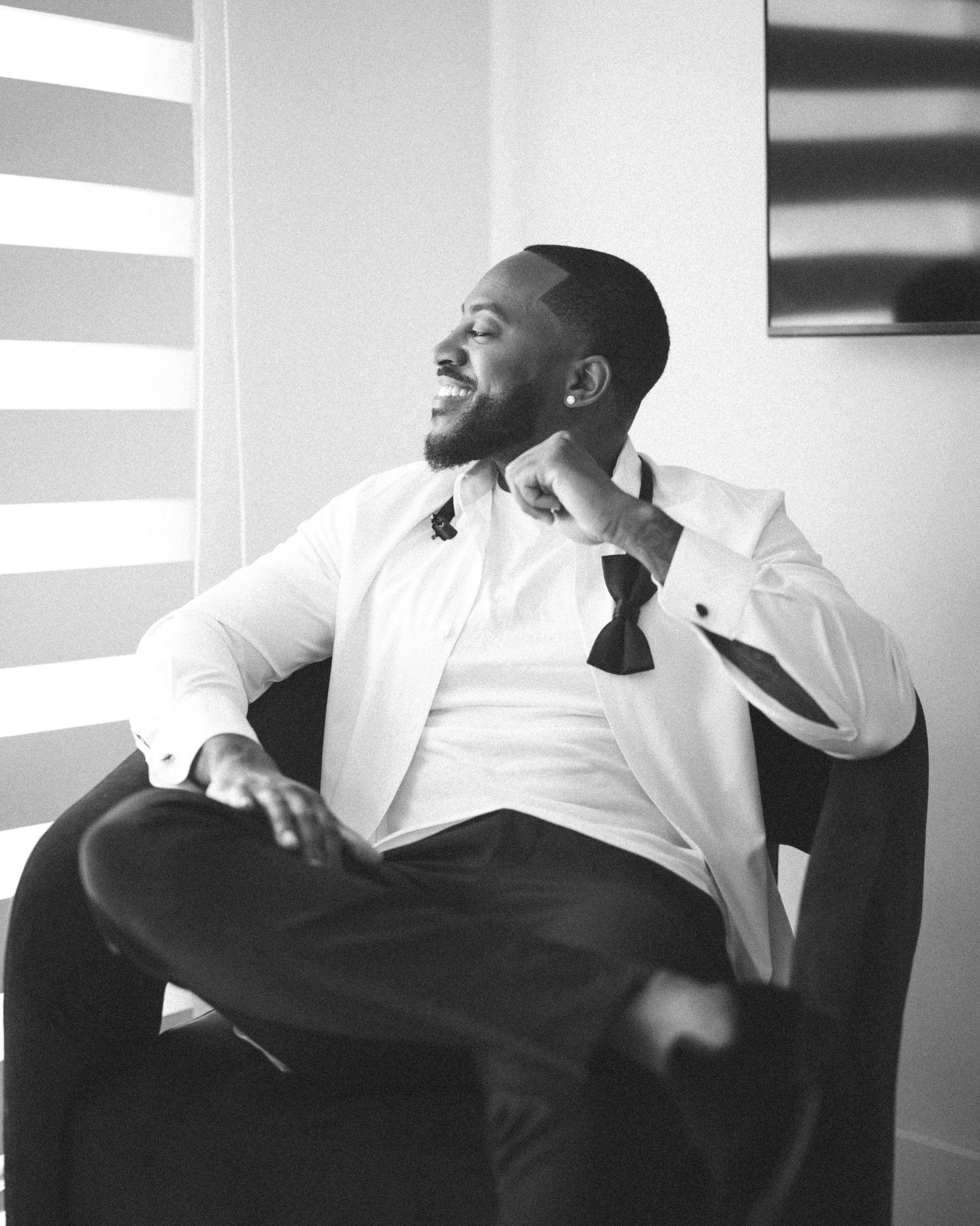 A well-dressed Black man in a white dress shirt and bow tie smiles while relaxing in a chair, black and white photo.