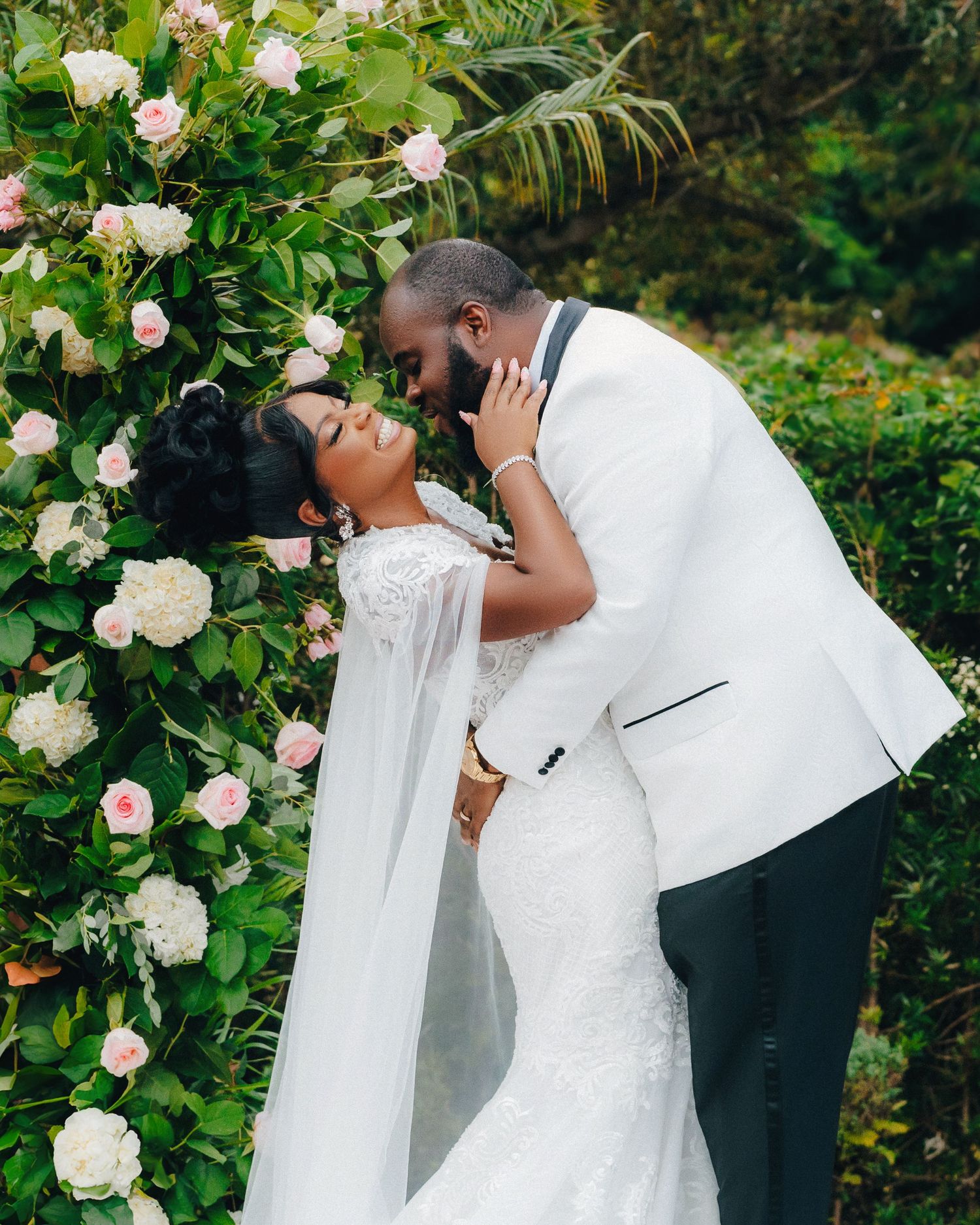 Elegant wedding couple poses against a backdrop of blooming roses in a garden setting.