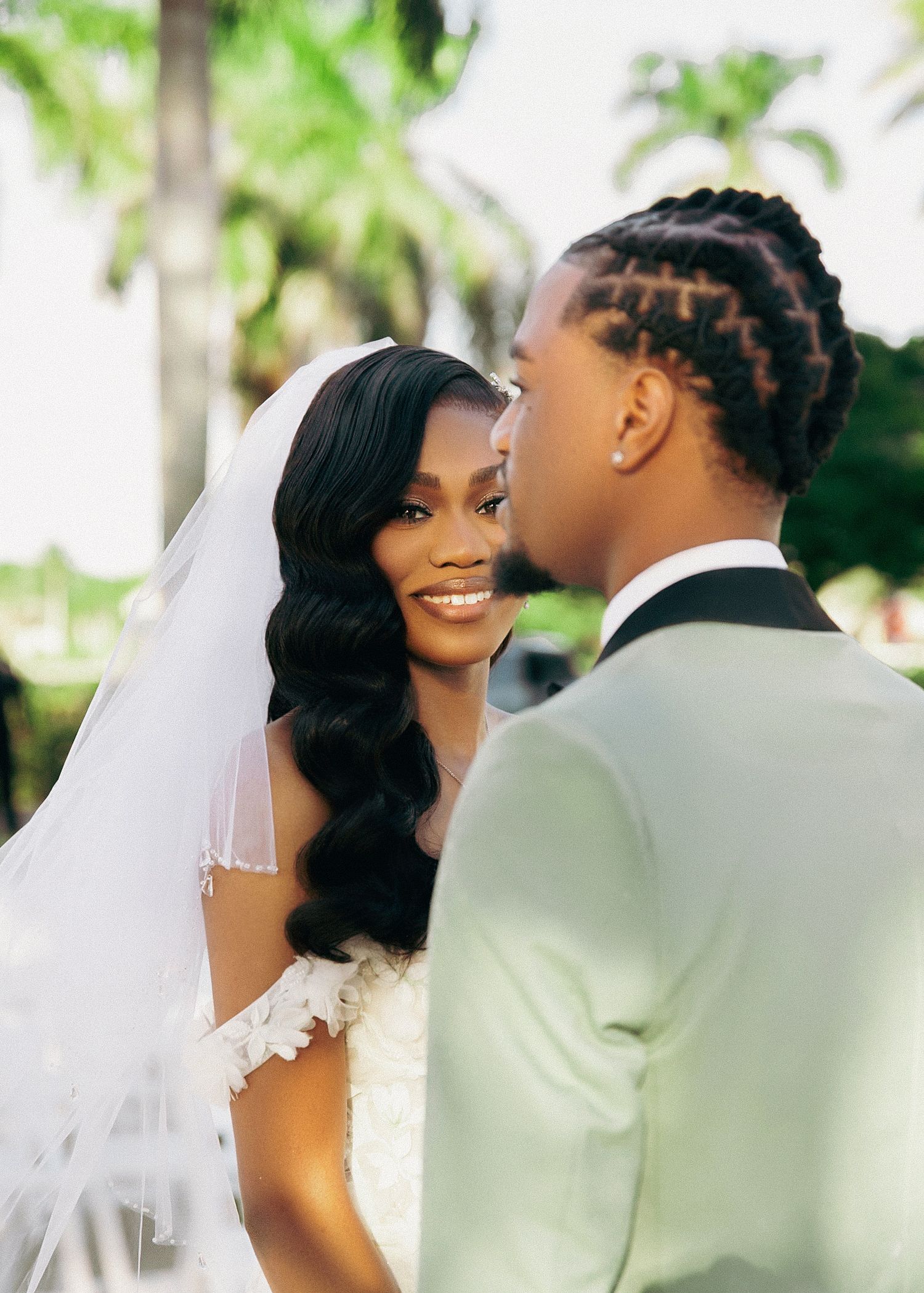 Romantic wedding moment captured outdoors with palm trees, featuring bride in flowing veil and soft waves hairstyle.
