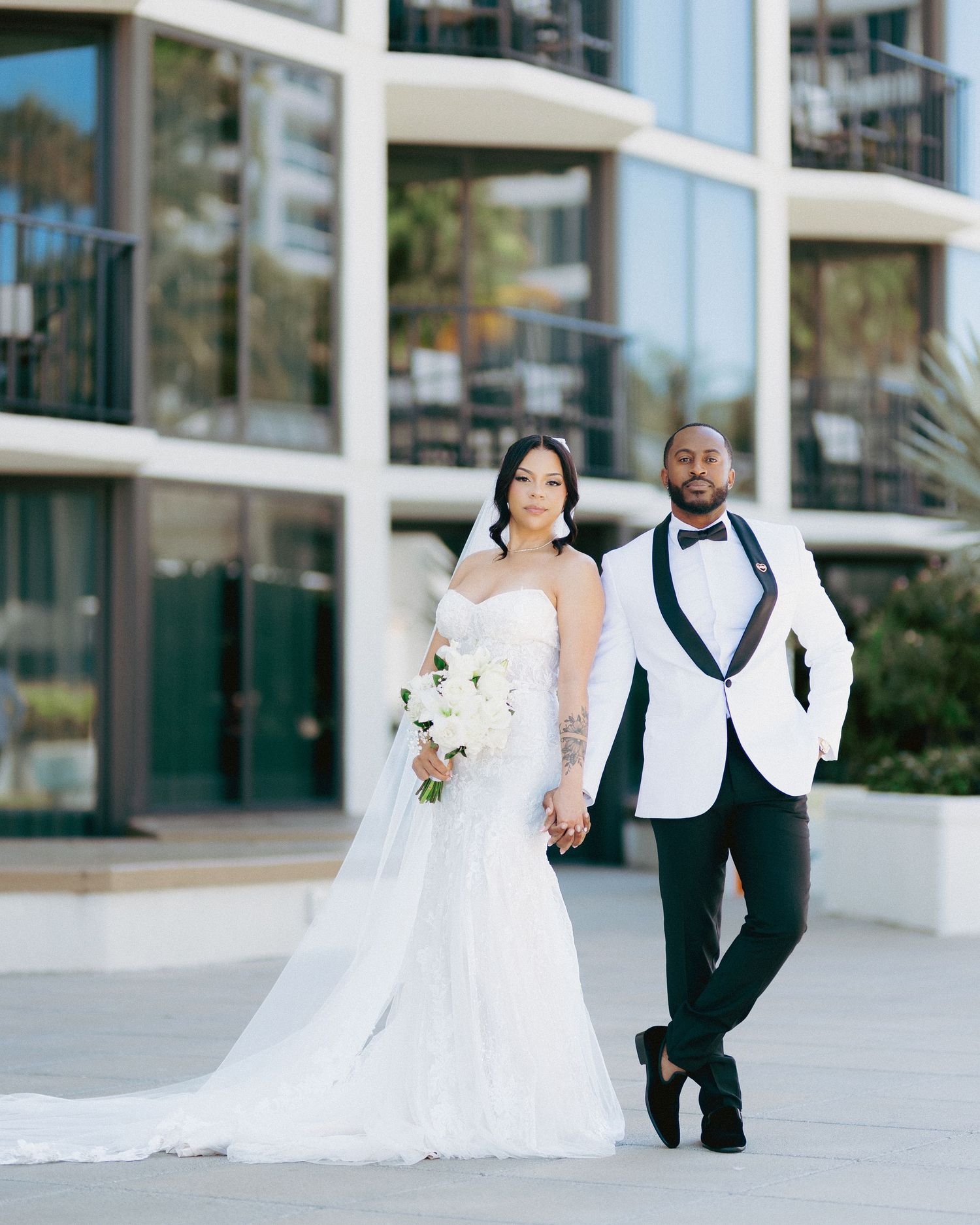 Elegant bride and groom pose outdoors in white wedding attire, she holds white bouquet, modern building backdrop.