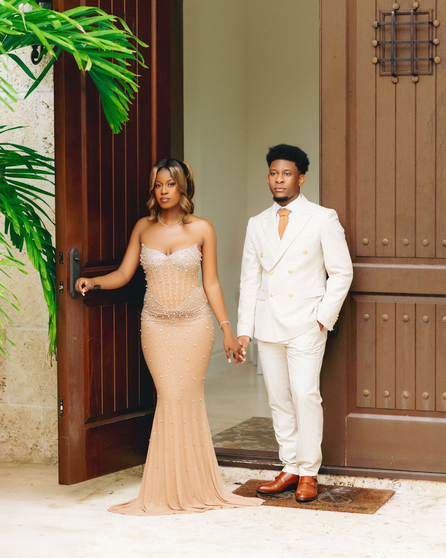 A couple in formal attire poses together against wooden doors, with the woman in a beige gown and man in white suit.