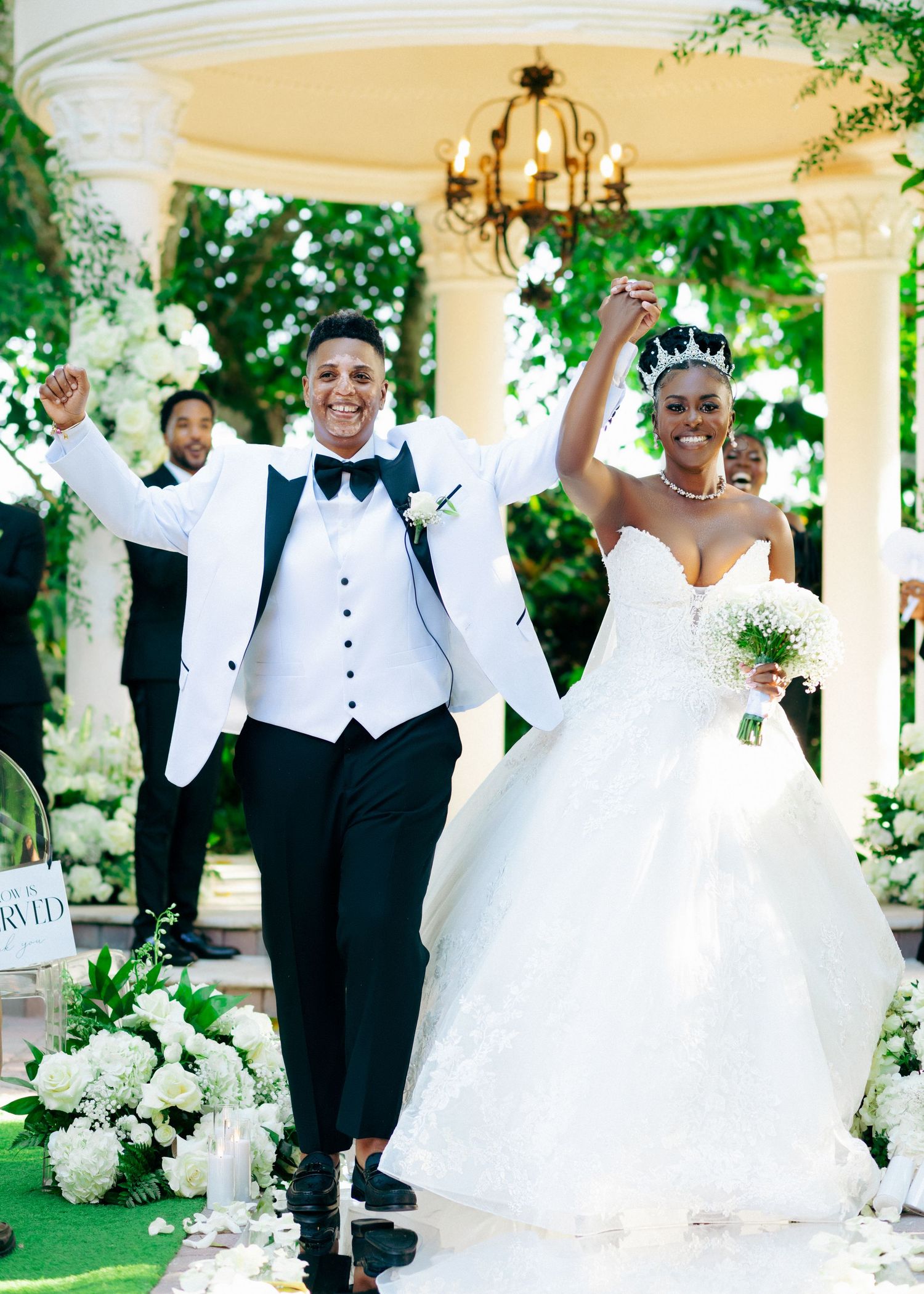 Wedding party celebrates joyfully under an elegant white gazebo surrounded by floral arrangements.
