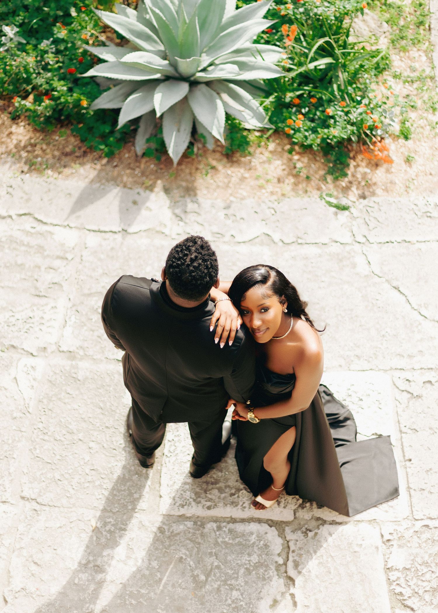 A couple in black attire sits together on stone steps surrounded by green plants and agave in an outdoor setting.