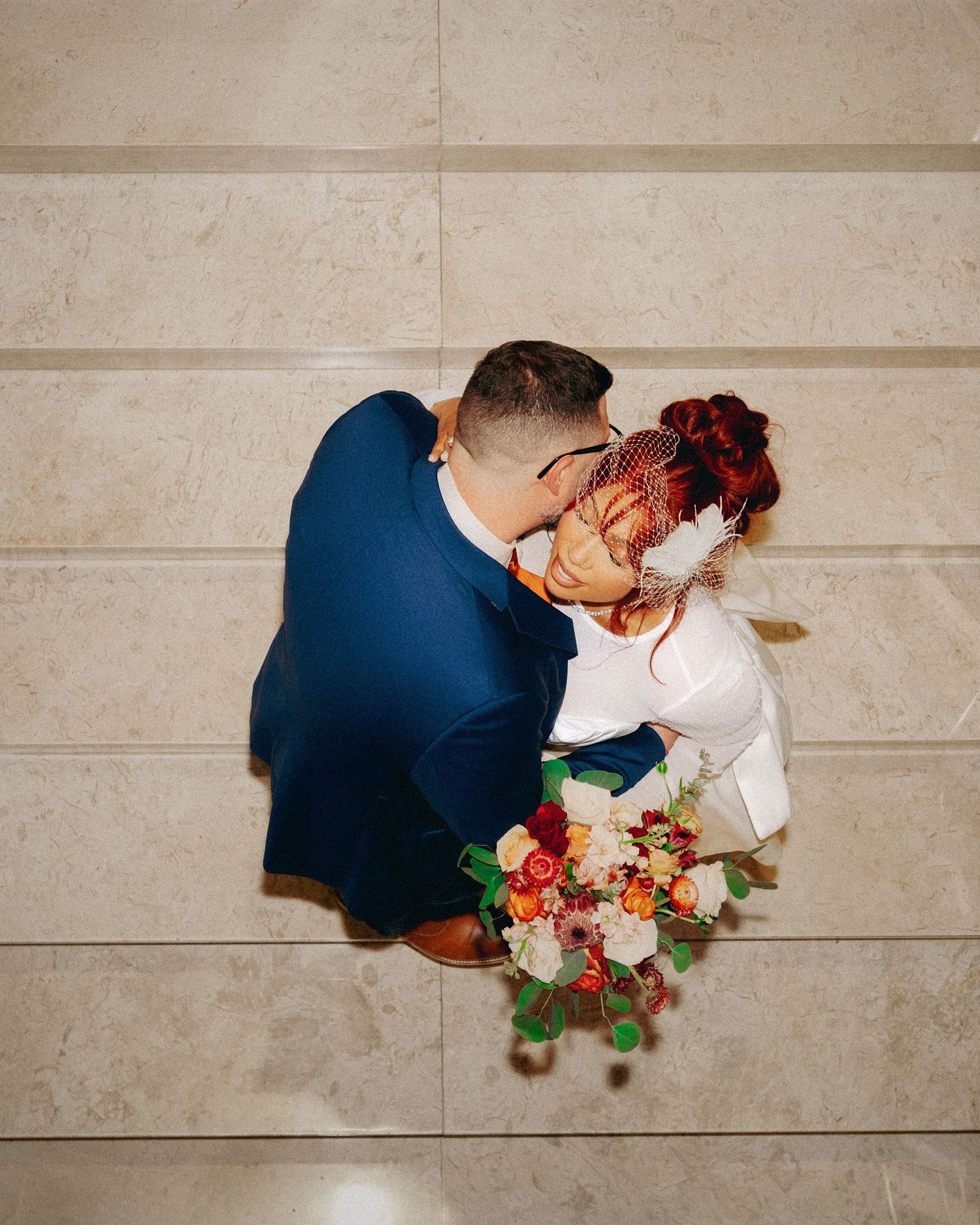 Aerial view of a couple sharing a tender moment on stone floor with vibrant red and white floral bouquet.