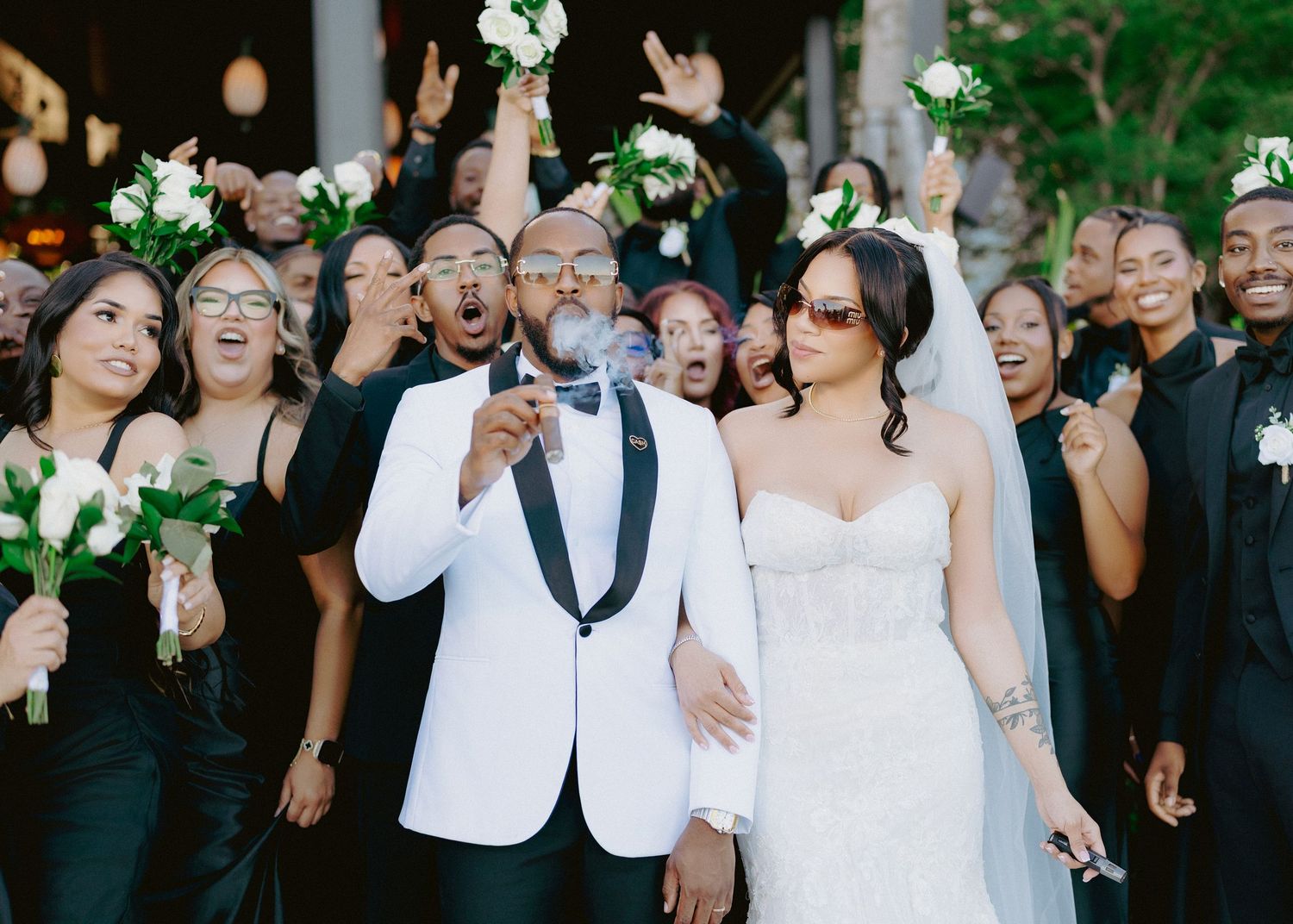 A joyful wedding party celebrates outdoors, bride in strapless gown and groom in white tuxedo surrounded by cheering guests.