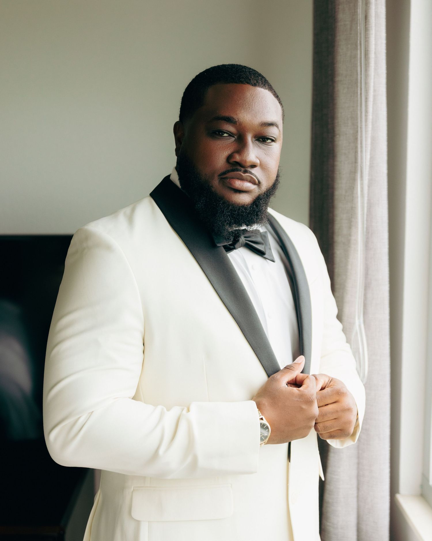 A groom in a classic white dinner jacket poses by a window.