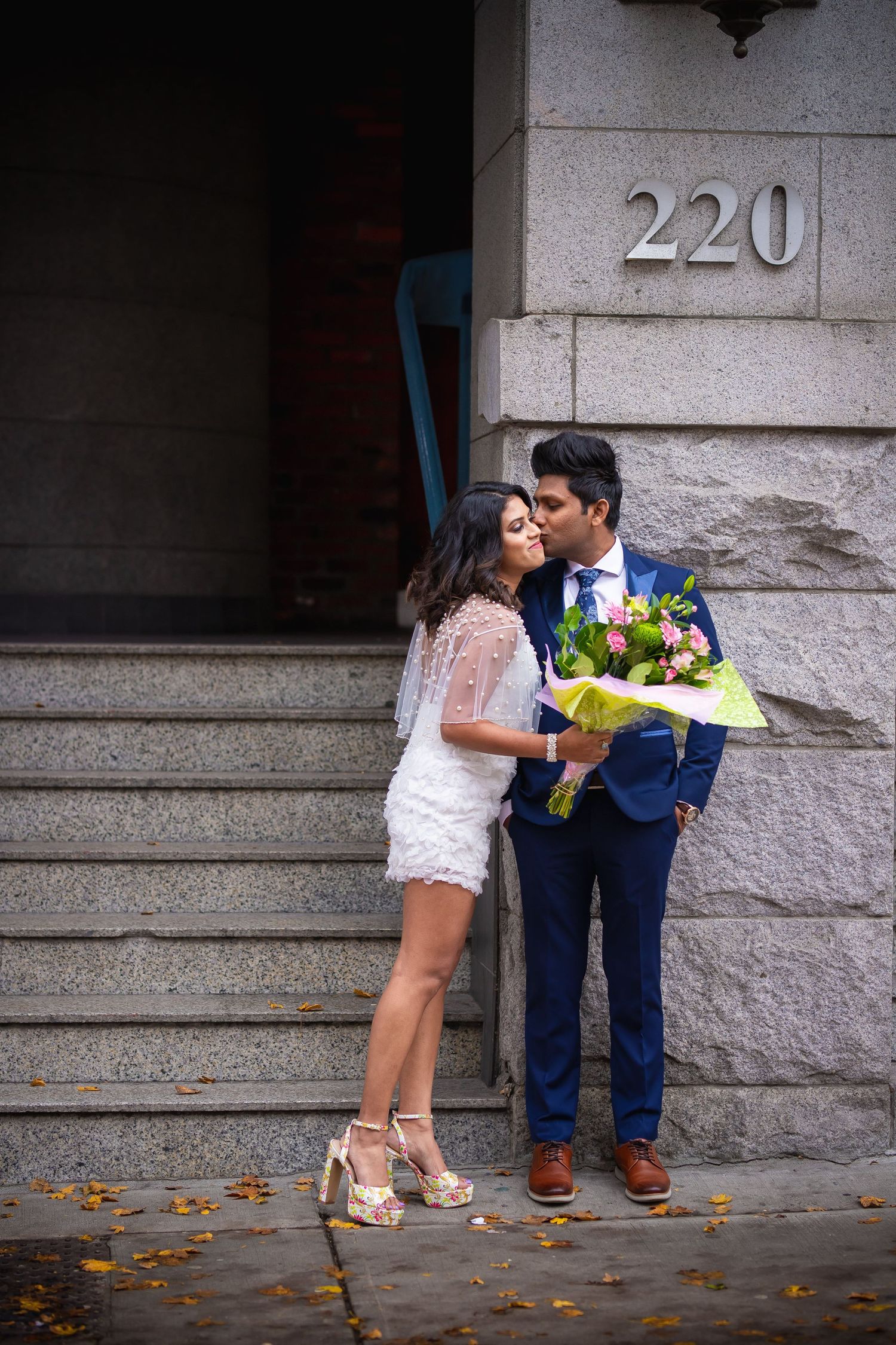 A couple in wedding attire shares a romantic kiss while holding colorful flowers on stone steps with autumn leaves.
