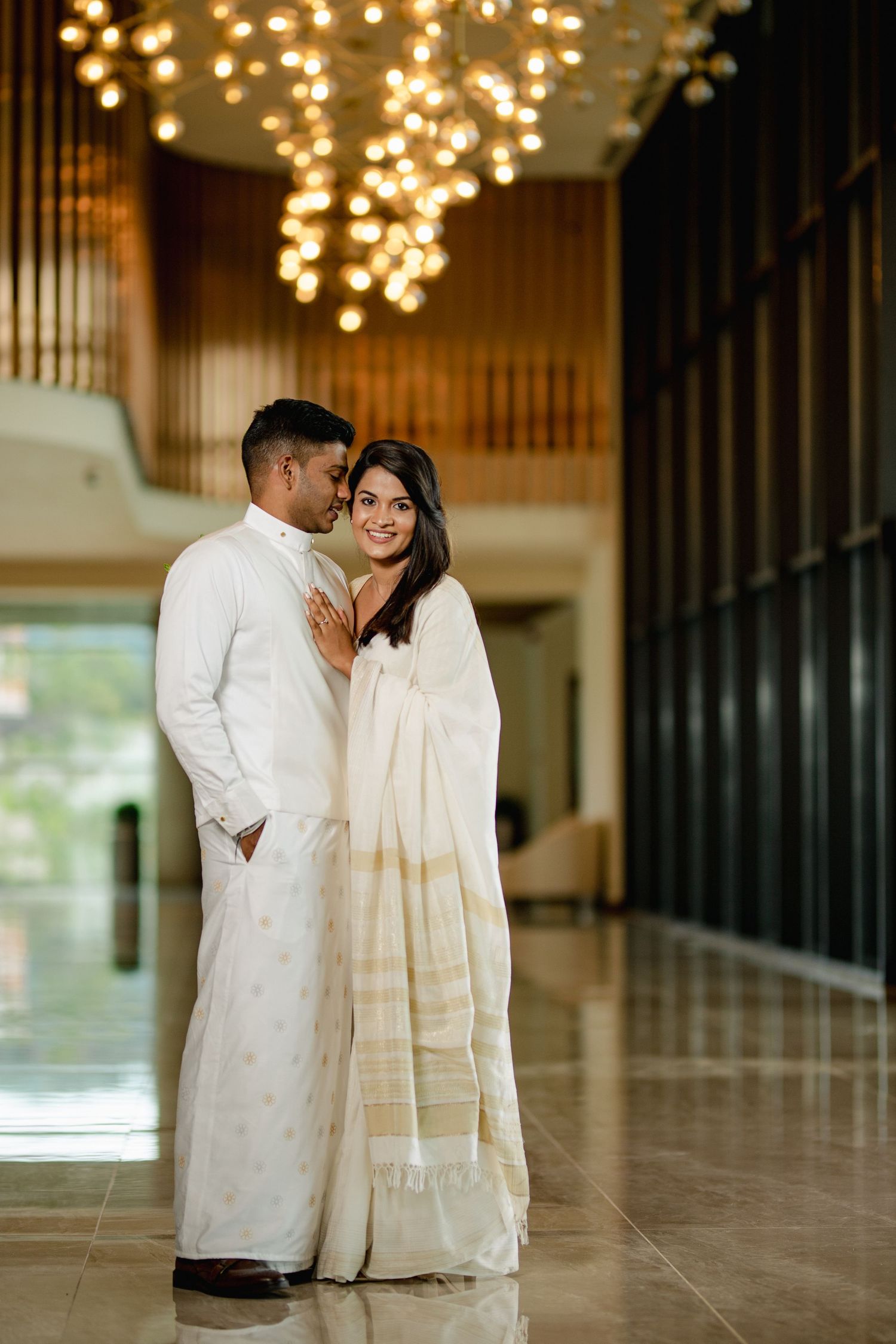 A couple in traditional white attire share an intimate moment in an elegant hallway with glowing chandeliers above.