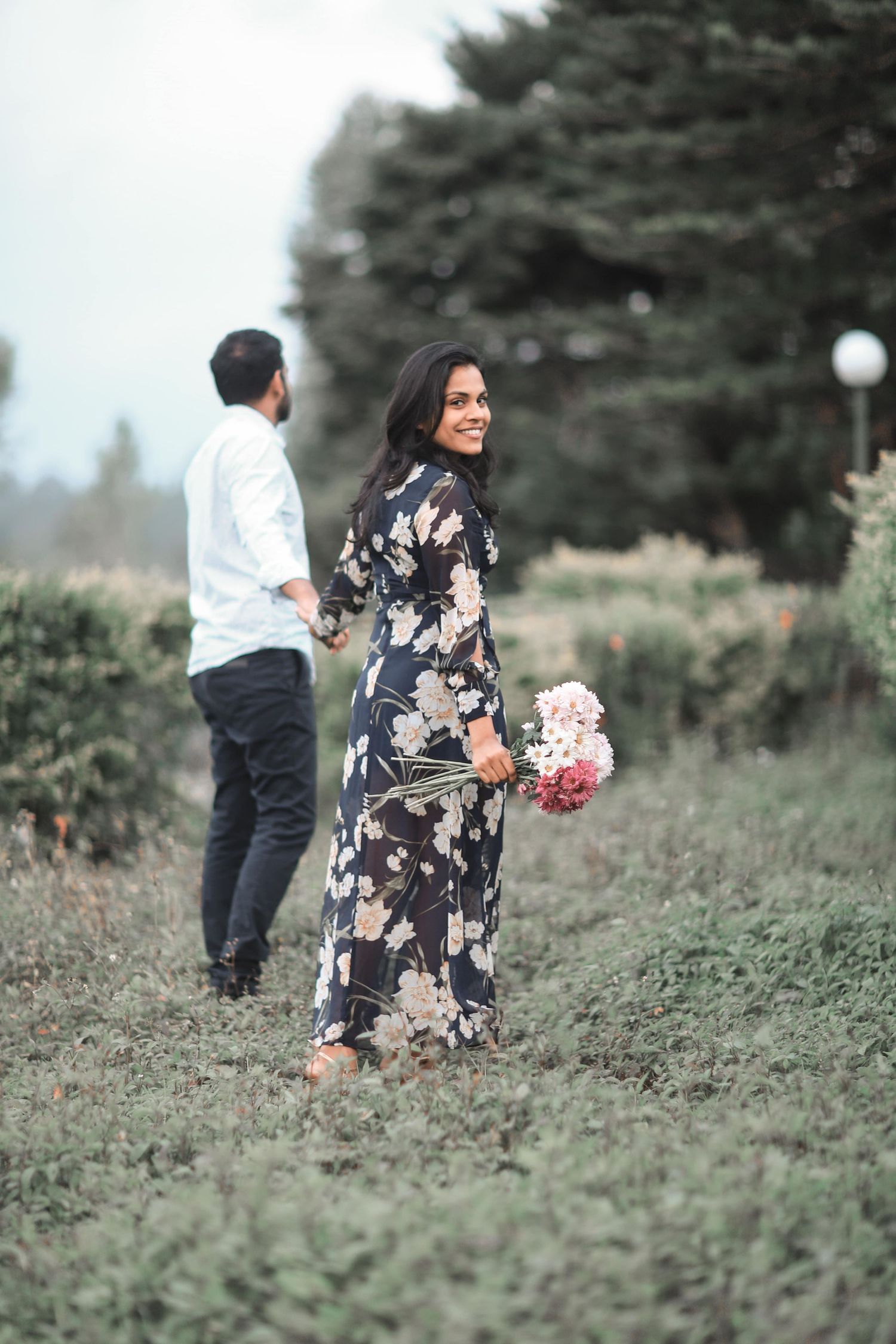 Couple in outdoor garden setting with floral dress and pink bouquet against evergreen trees in background.