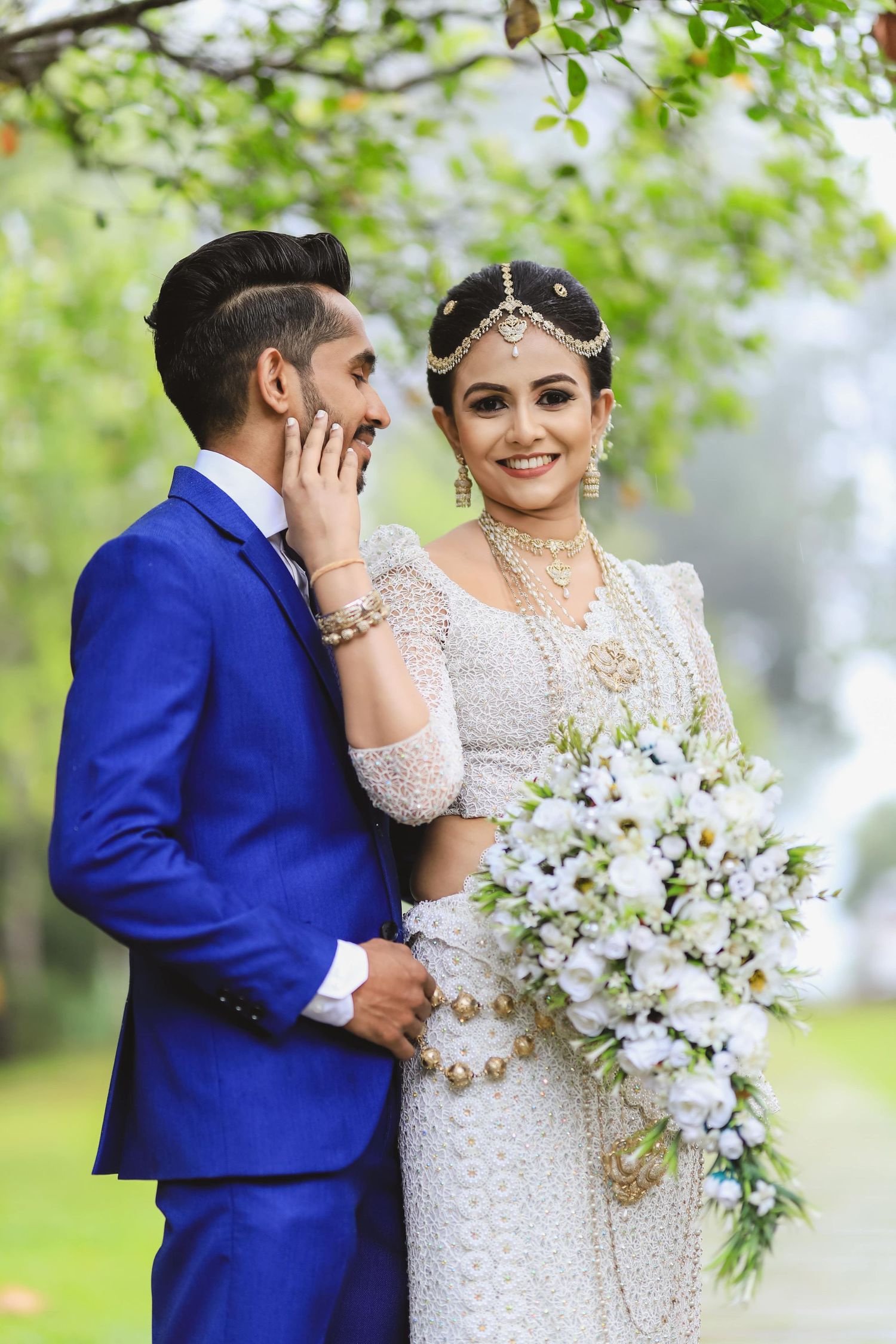 A wedding couple in royal blue suit and white lace dress pose together with a white floral bouquet in a garden setting.