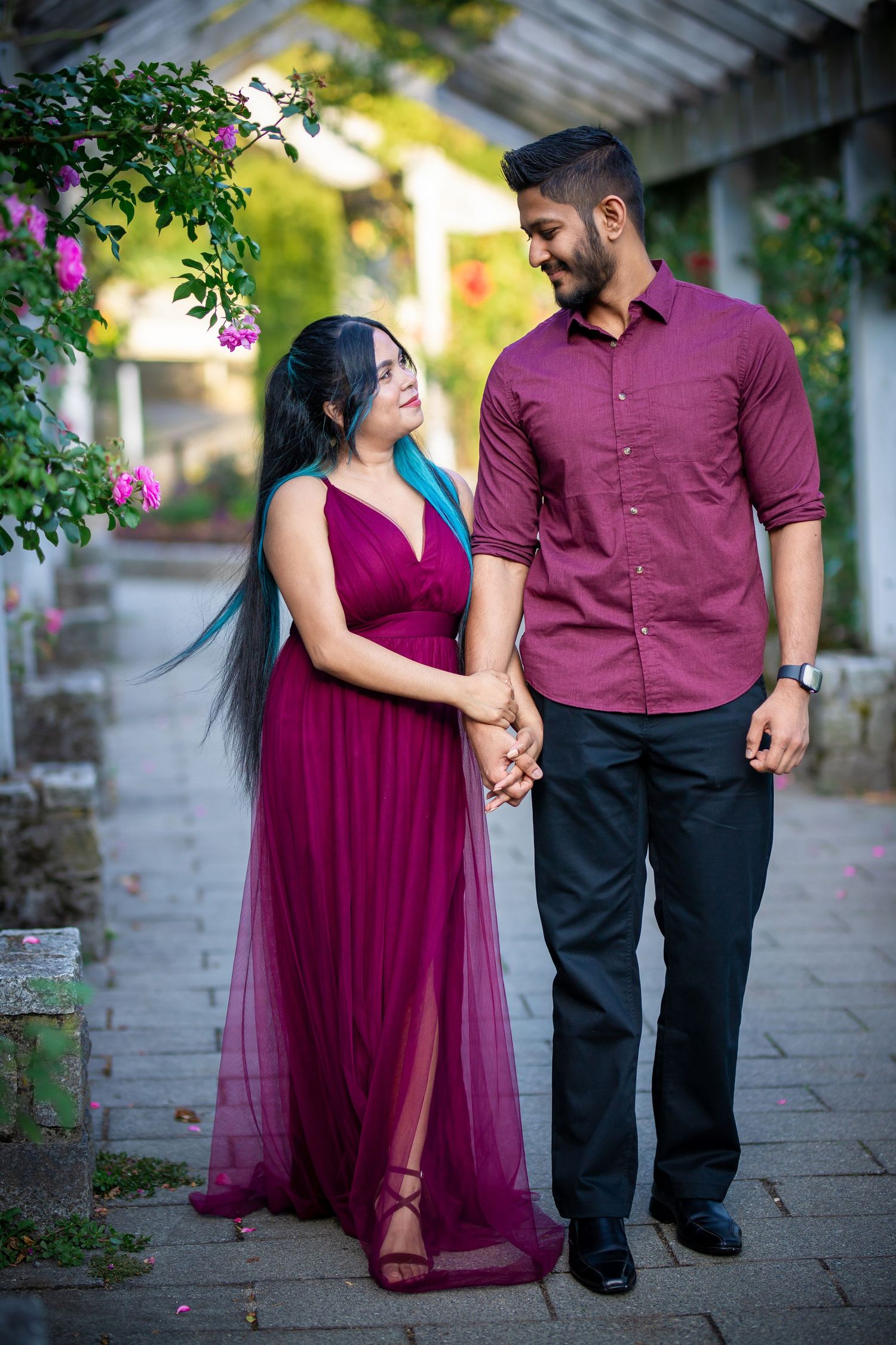 A couple in coordinating burgundy outfits hold hands while walking along a garden path during a romantic photoshoot.