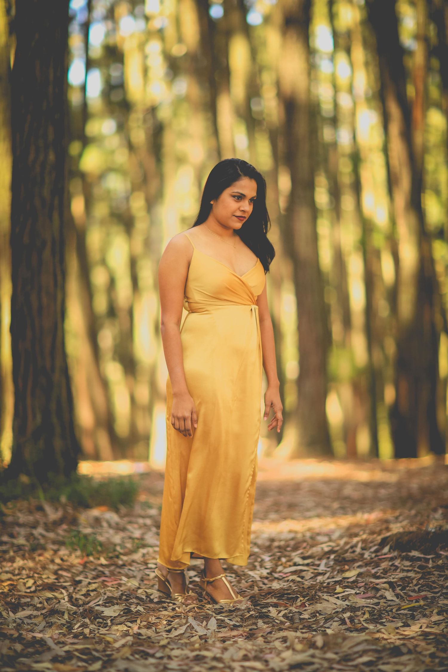 A woman in a flowing yellow dress poses gracefully in a sunlit forest setting during a maternity photoshoot.