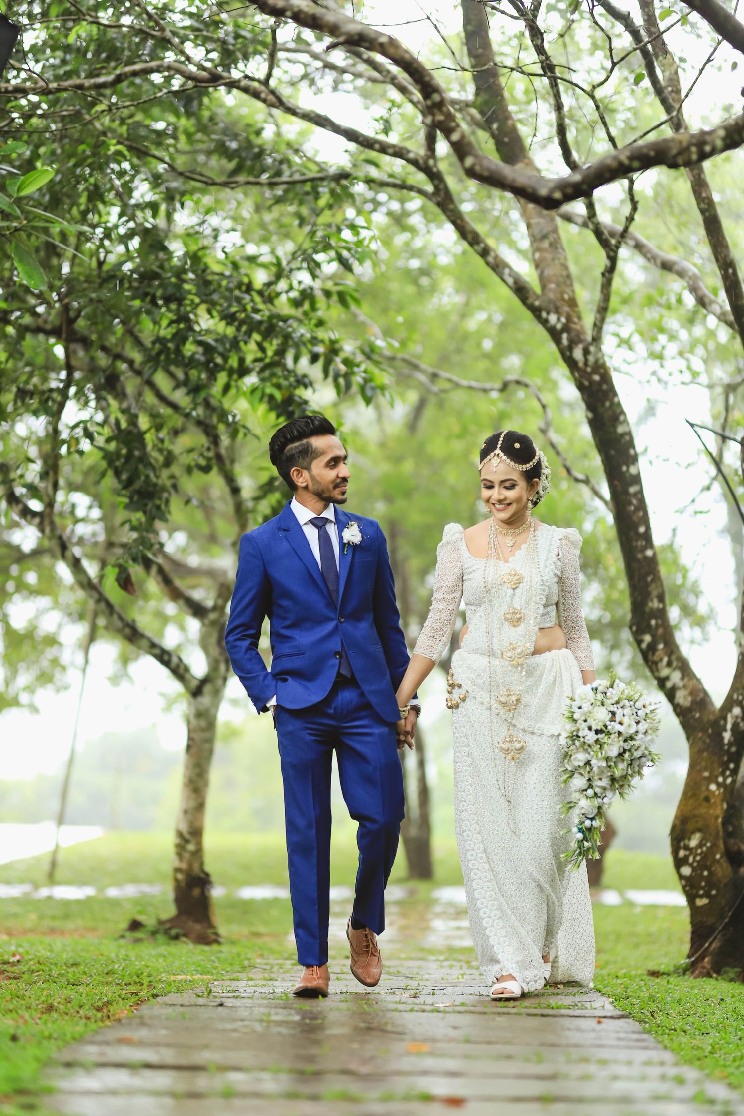 A couple walking along a tree-lined path in elegant wedding attire.