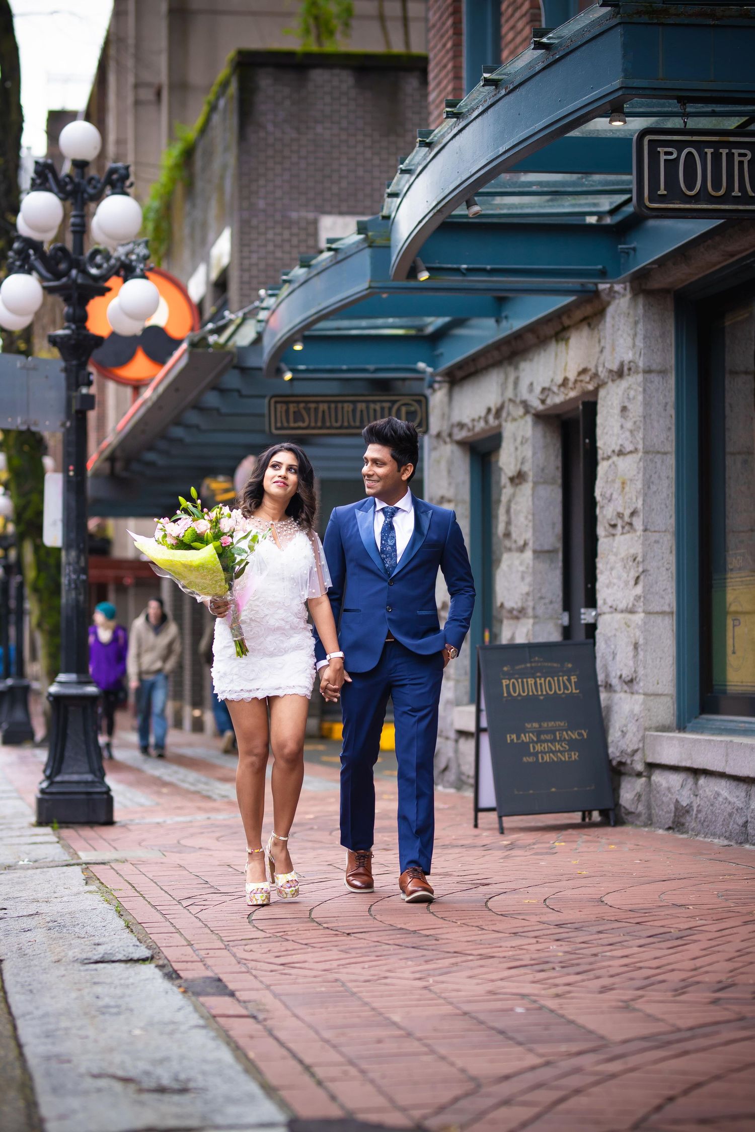A couple walks down a charming brick sidewalk in front of historic storefronts with blue awnings.