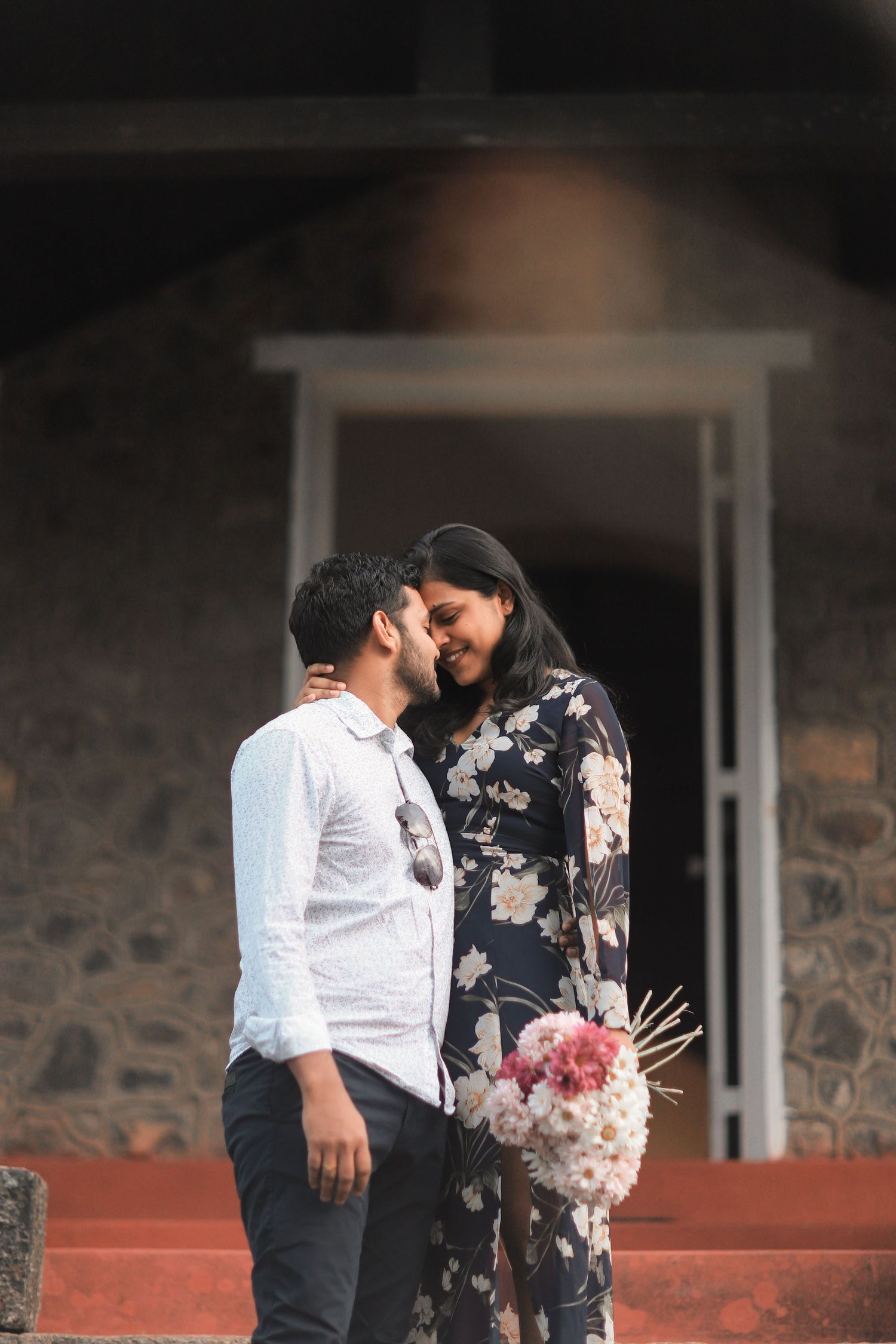 A couple in floral dress and white shirt share an intimate moment against a stone wall backdrop.