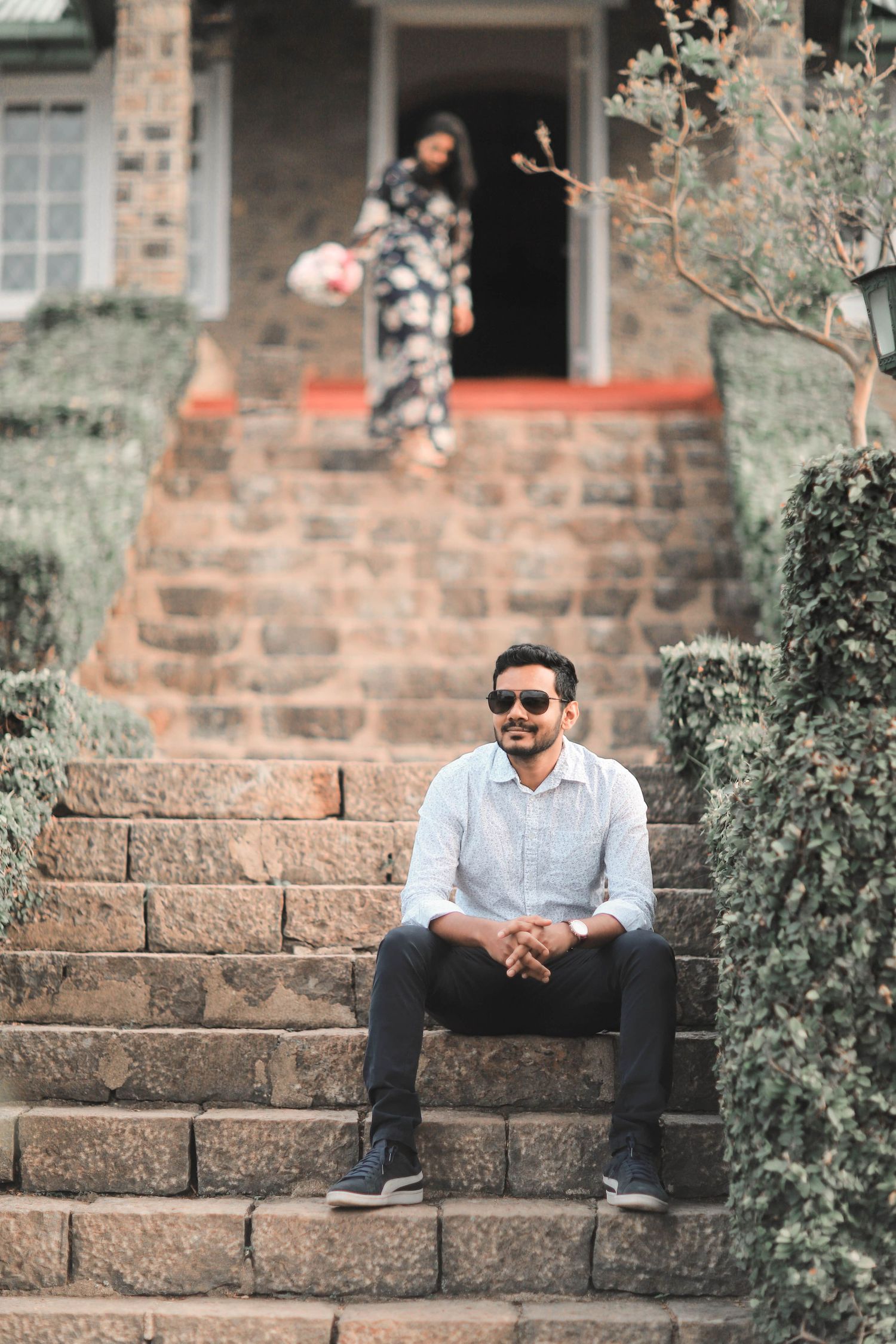 Person in sunglasses sits casually on brick steps surrounded by greenery in an outdoor setting.