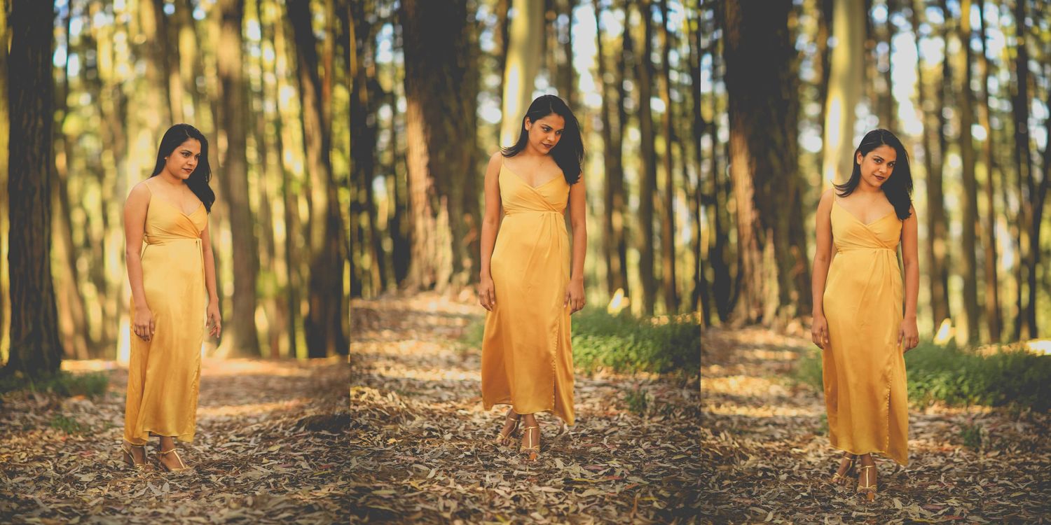 A person in a flowing yellow dress stands in a sunlit forest path surrounded by tall pine trees during golden hour.