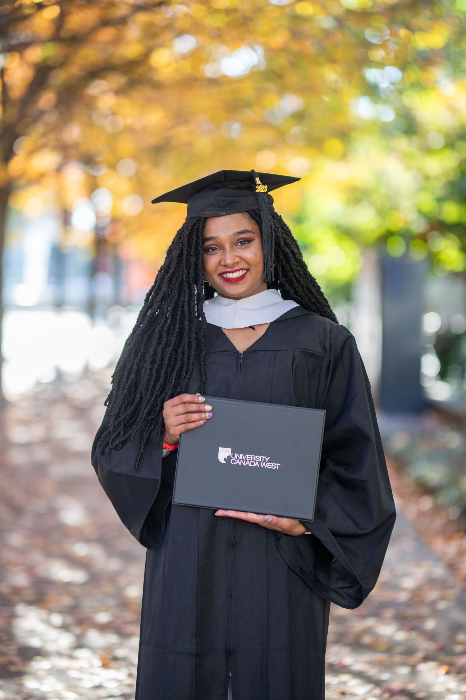 Graduate in traditional black cap and gown poses with diploma among fall foliage.
