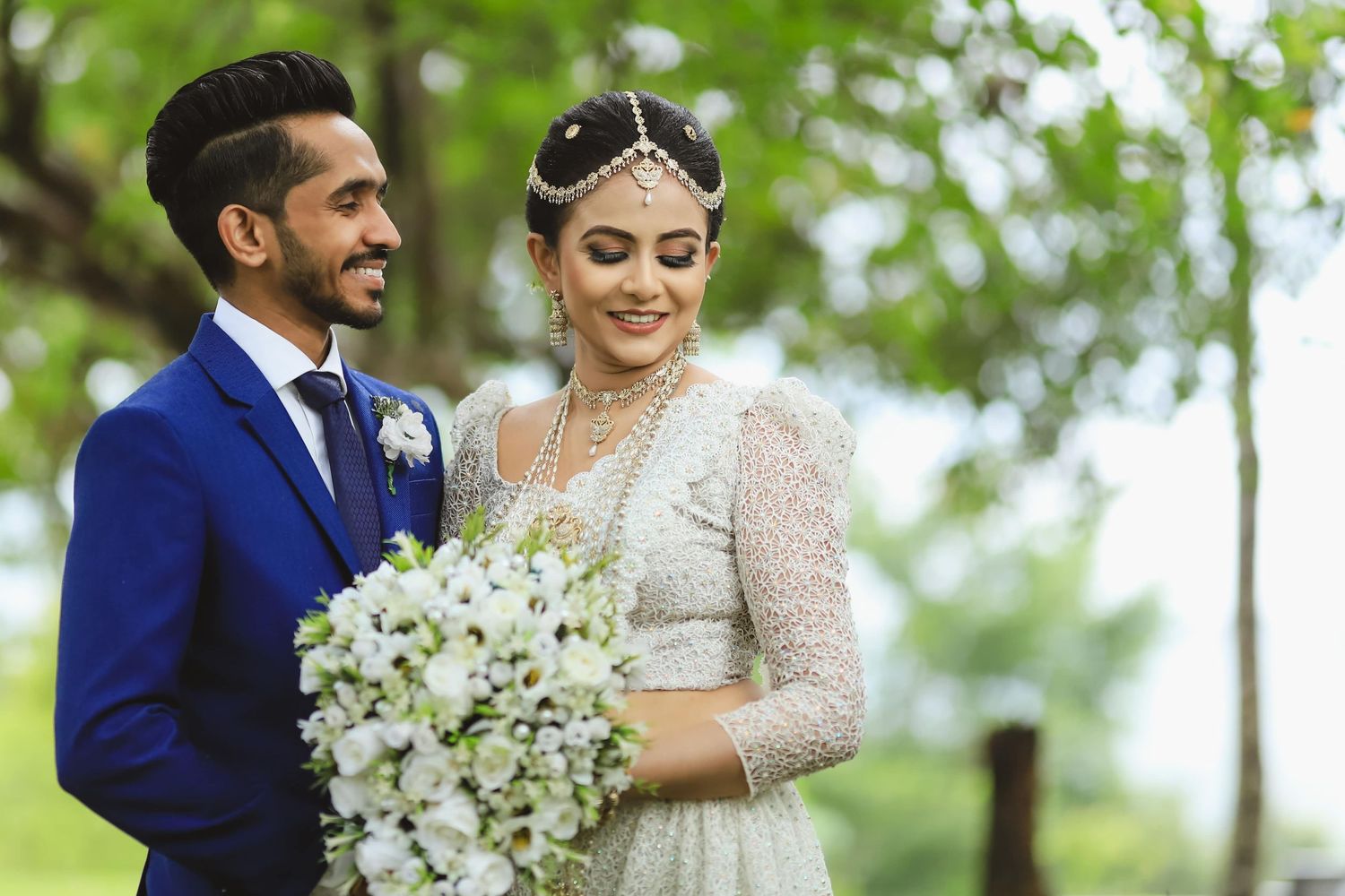 A couple in traditional South Asian wedding attire posing outdoors with white floral bouquet.