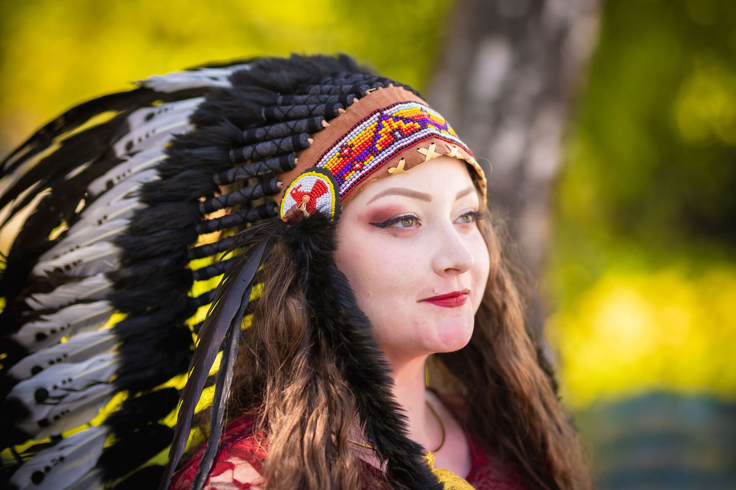 A person wearing a Native American headdress poses outdoors against a blurred green background.