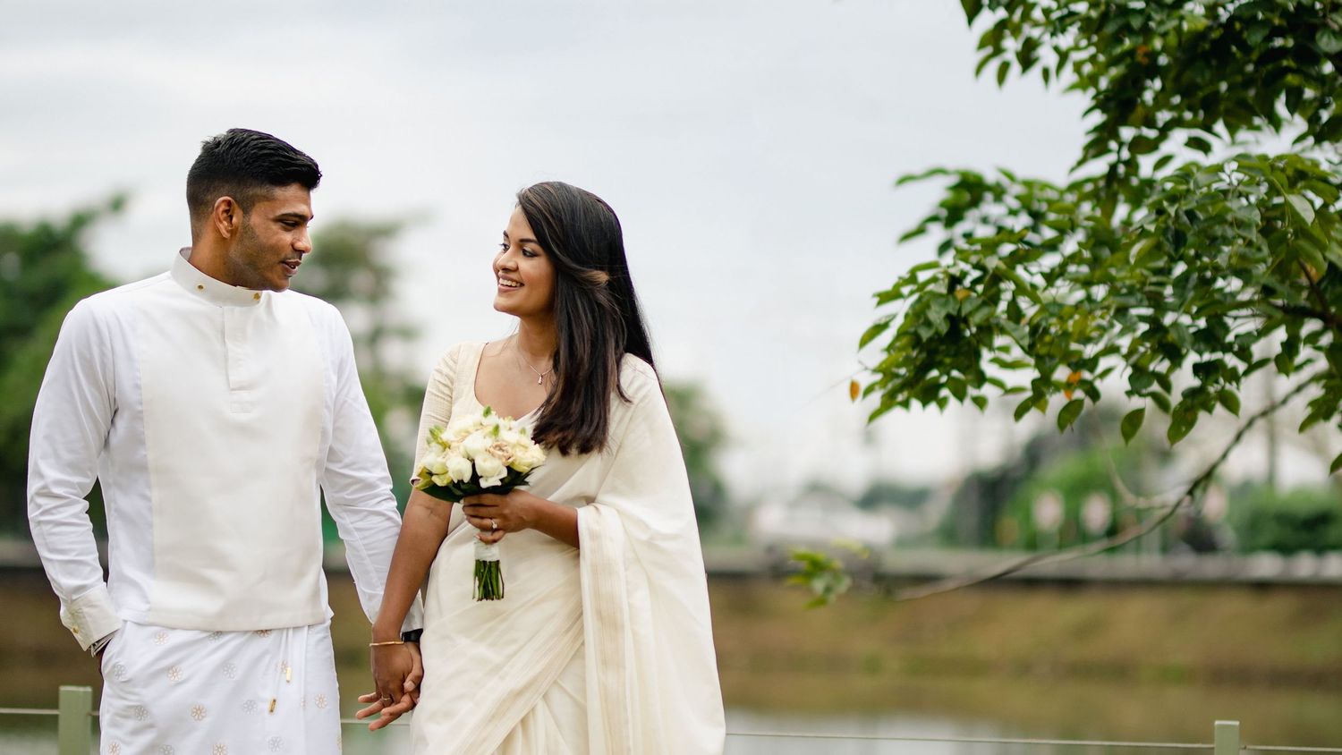 A couple in traditional wedding attire walks together in a lush green garden setting on their special day.